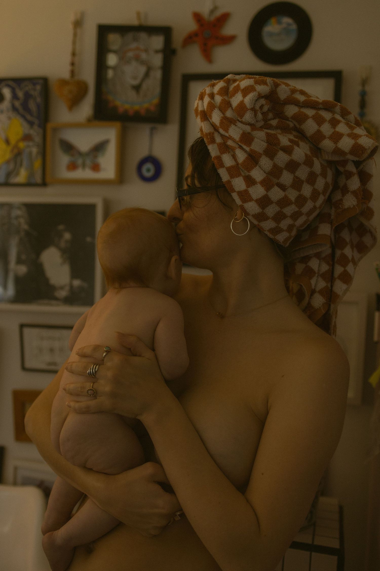 A mother with a checkered towel wrap kisses her newborn baby, surrounded by eclectic wall art in a warm home.