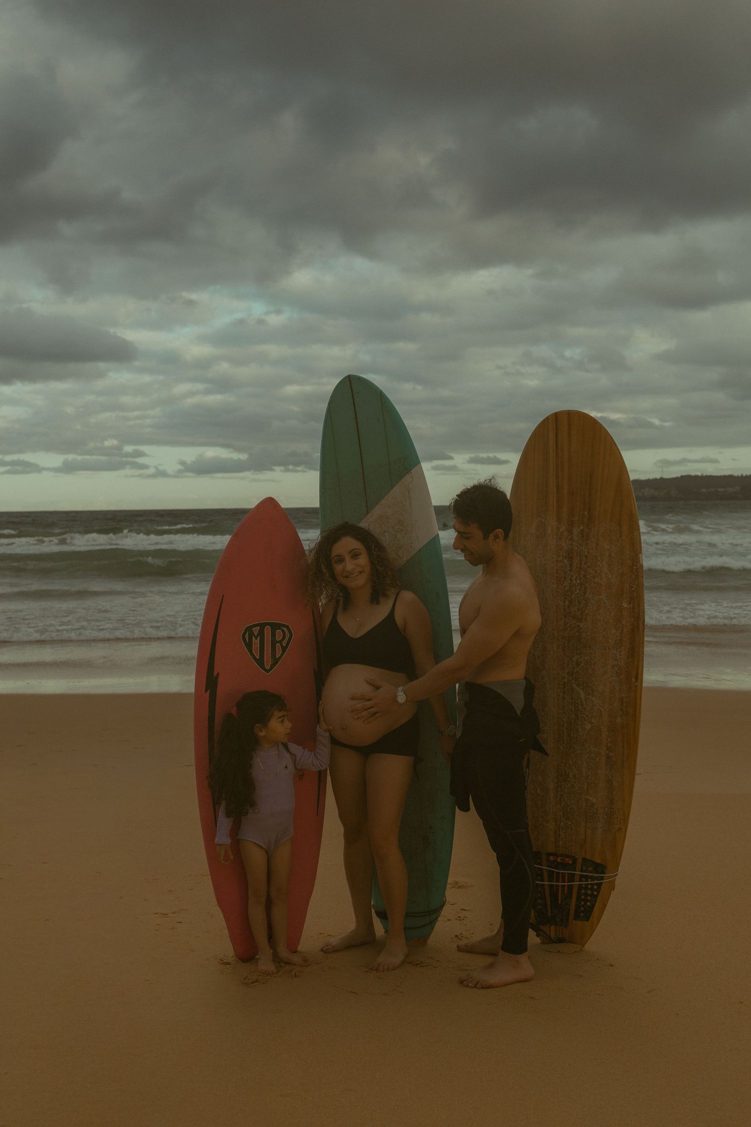 Pregnant woman with family and surfboards on a cloudy beach, young girl and man touching her baby bump.