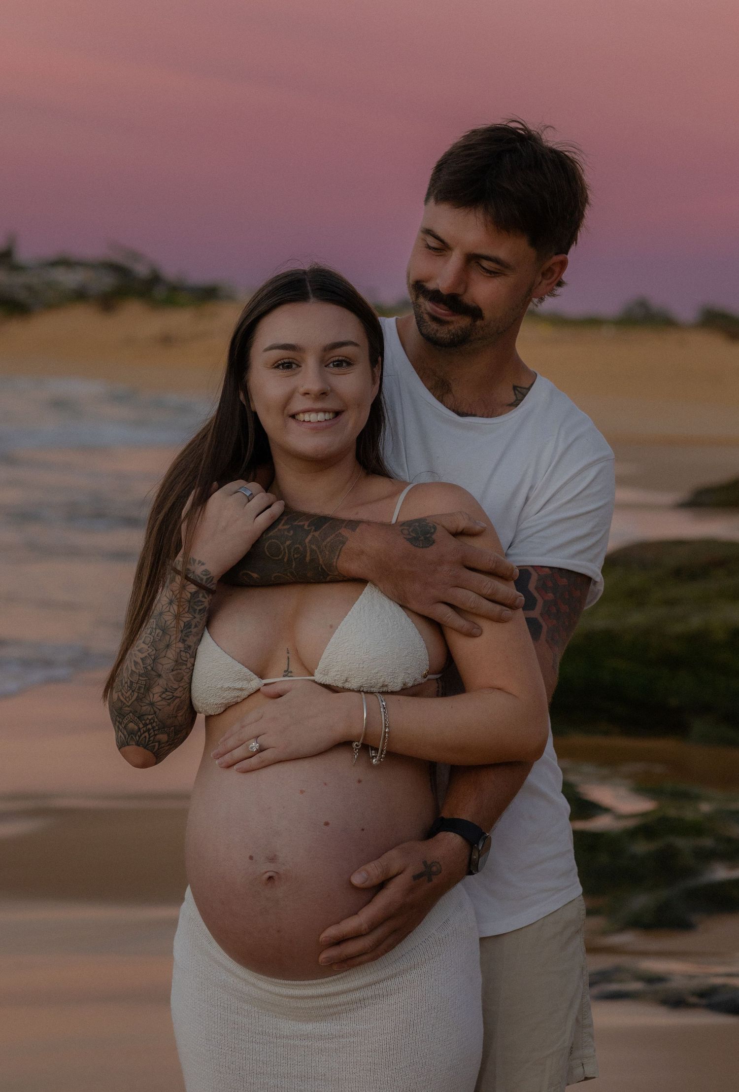 Man embracing pregnant woman from behind on beach at sunset, both smiling, showing baby bump.