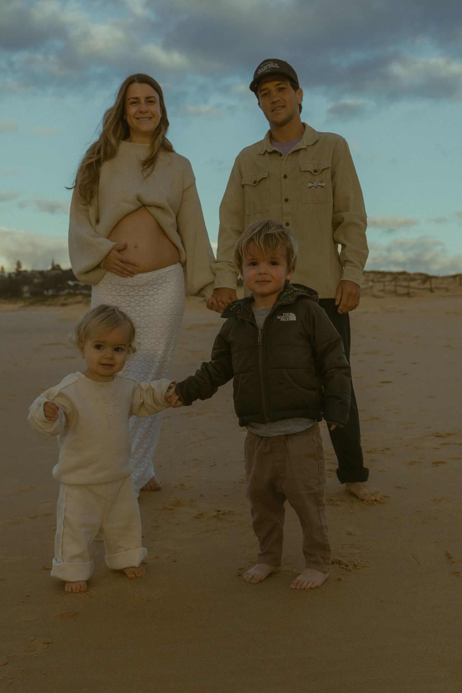 Family of four poses on sandy beach at sunset, pregnant mother and father stand with two young barefoot children.