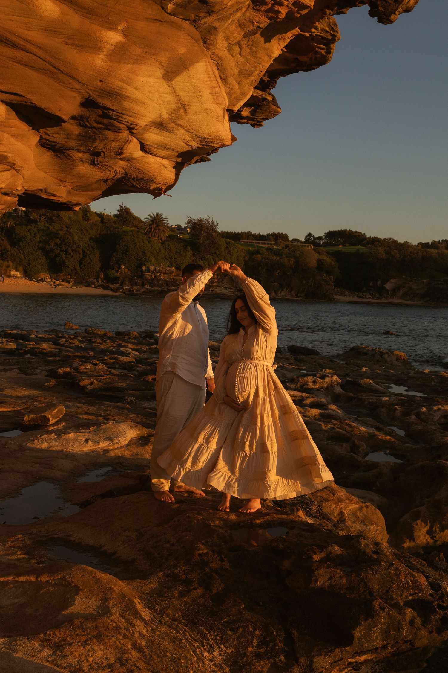Couple in white outfits dancing on rocky shore at golden sunset beneath sandstone overhang near calm water.