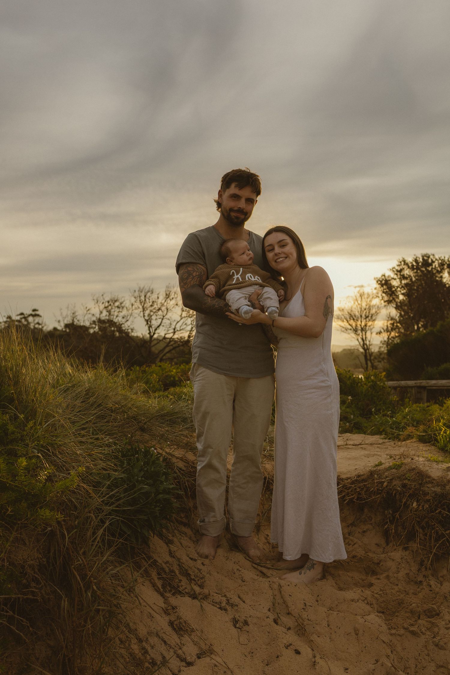 A couple holding their baby poses on a sandy path at sunset, surrounded by green grass and a dramatic sky.
