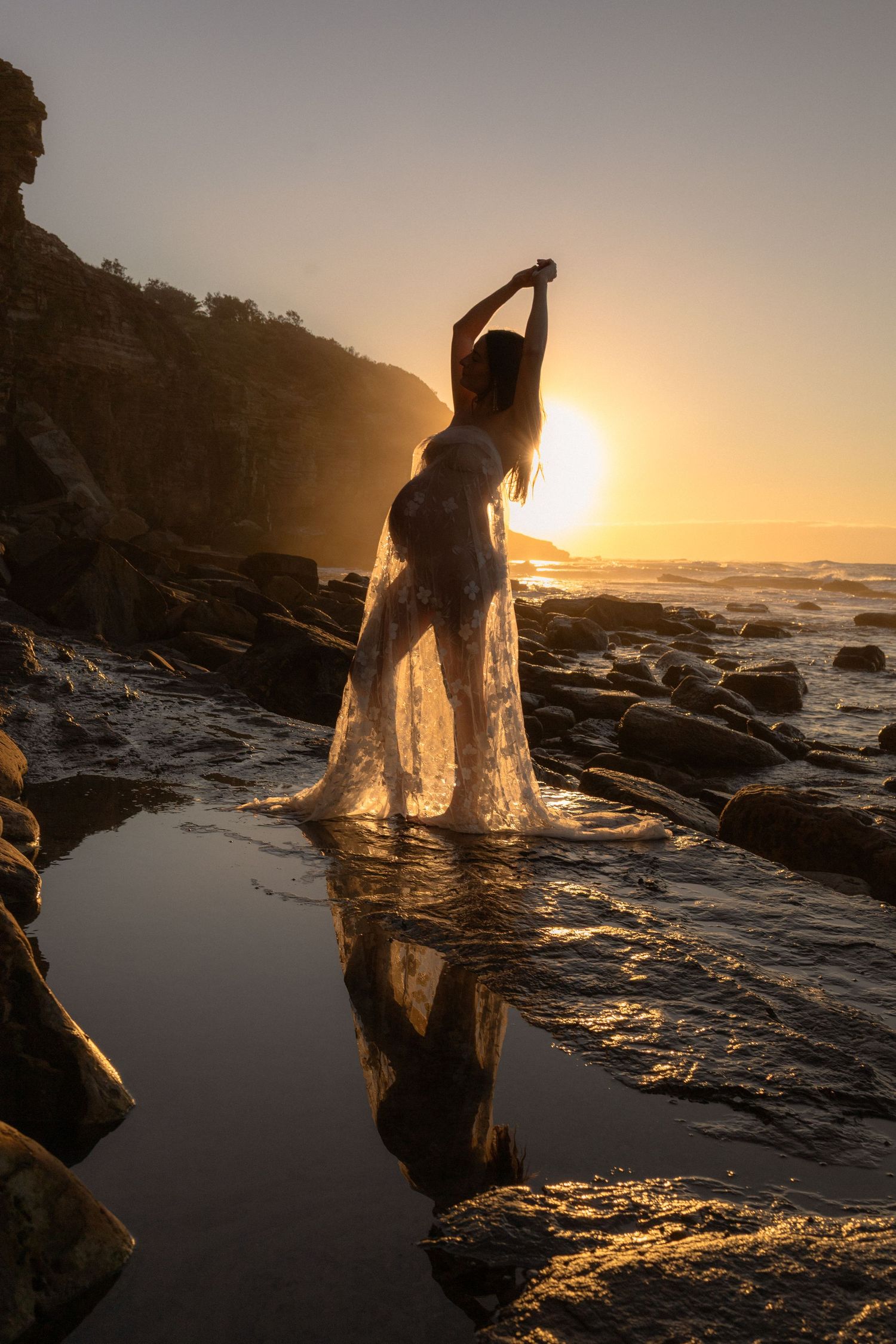Silhouette of a woman in flowing white dress on rocky beach at golden sunset with ocean reflection.