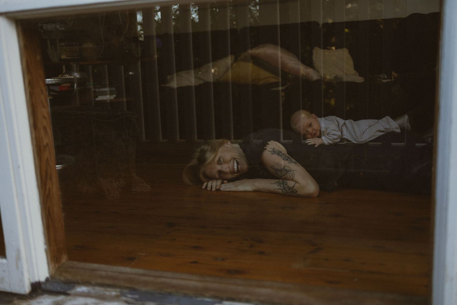 Two children lying on a hardwood floor inside a home, viewed through a glass sliding door.