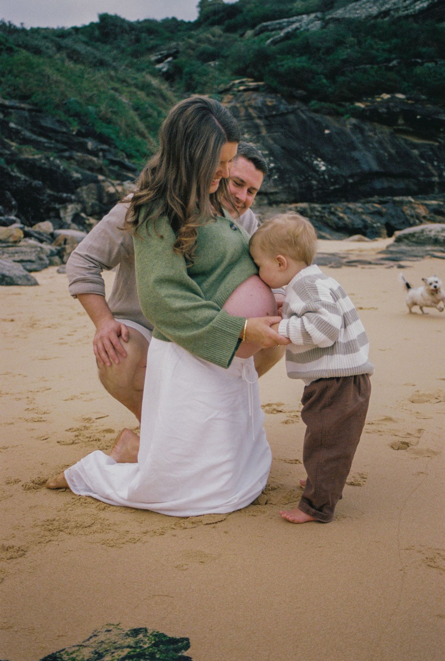 Pregnant woman kneeling on beach with toddler and partner, rocky coastal backdrop, family maternity photo session.