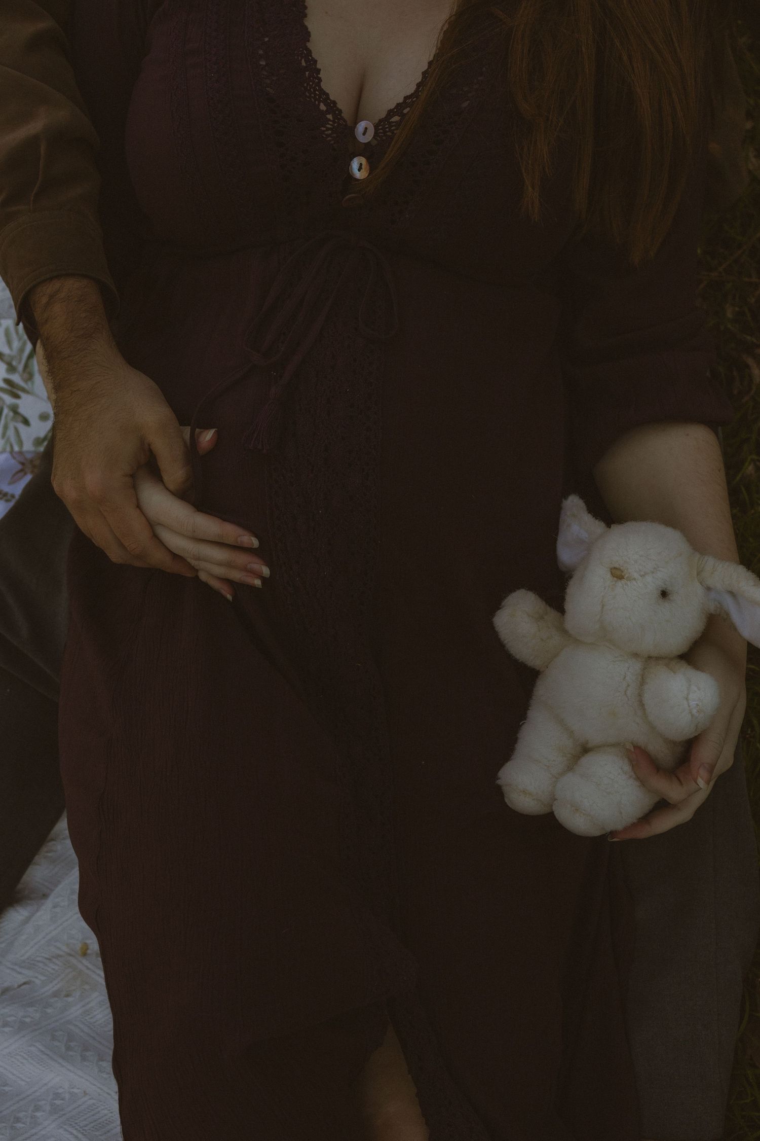 Pregnant woman in dark dress holding white stuffed bunny toy while partner gently touches her baby bump.