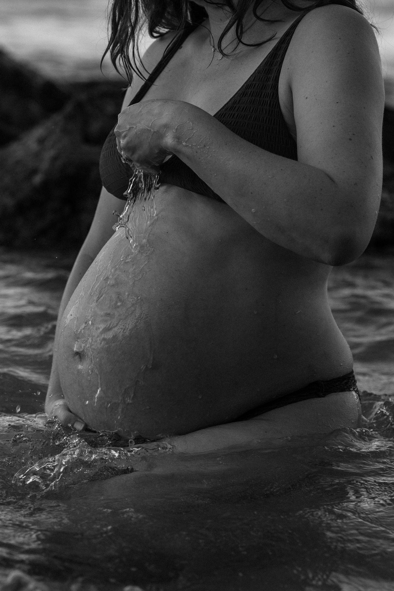 Black and white photo of a pregnant woman's torso in water with droplets cascading down her belly.