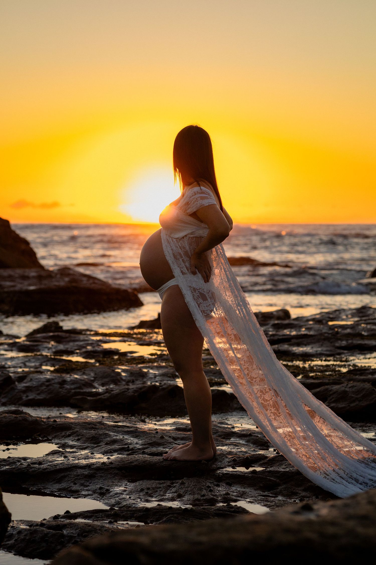 Pregnant woman in white lace wrap stands on rocky beach at golden sunset with ocean backdrop.