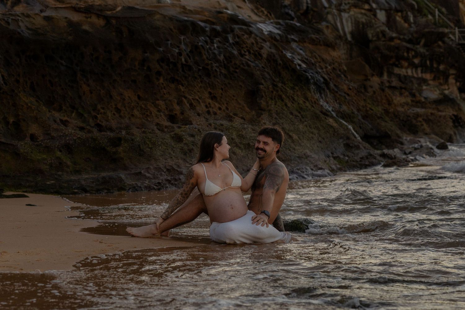 Couple sitting together in shallow ocean water on a rocky beach, woman in white bikini with baby bump, man with tattoos.