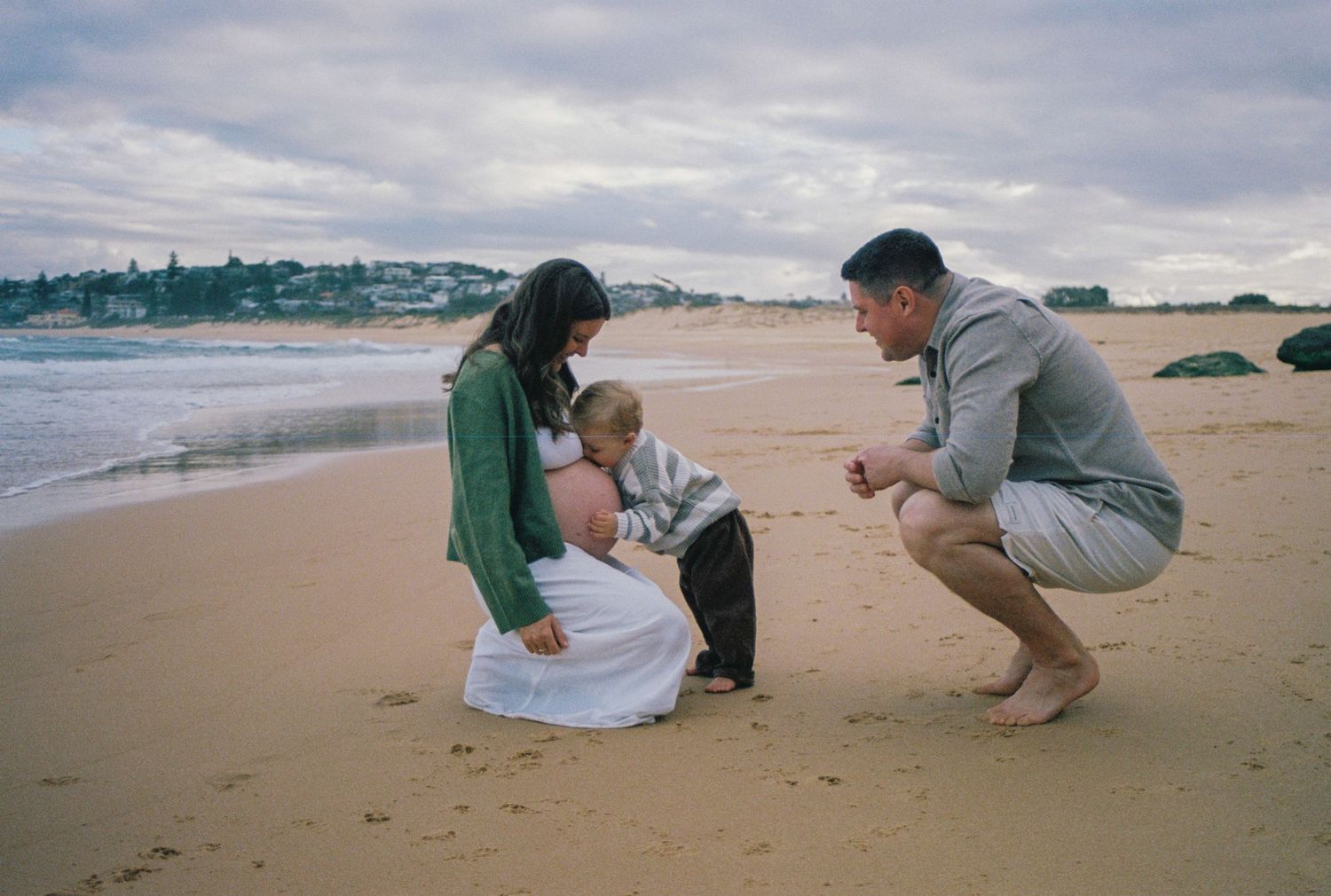 Family of three sharing a tender moment on a sandy beach, with waves and cloudy skies in the background.