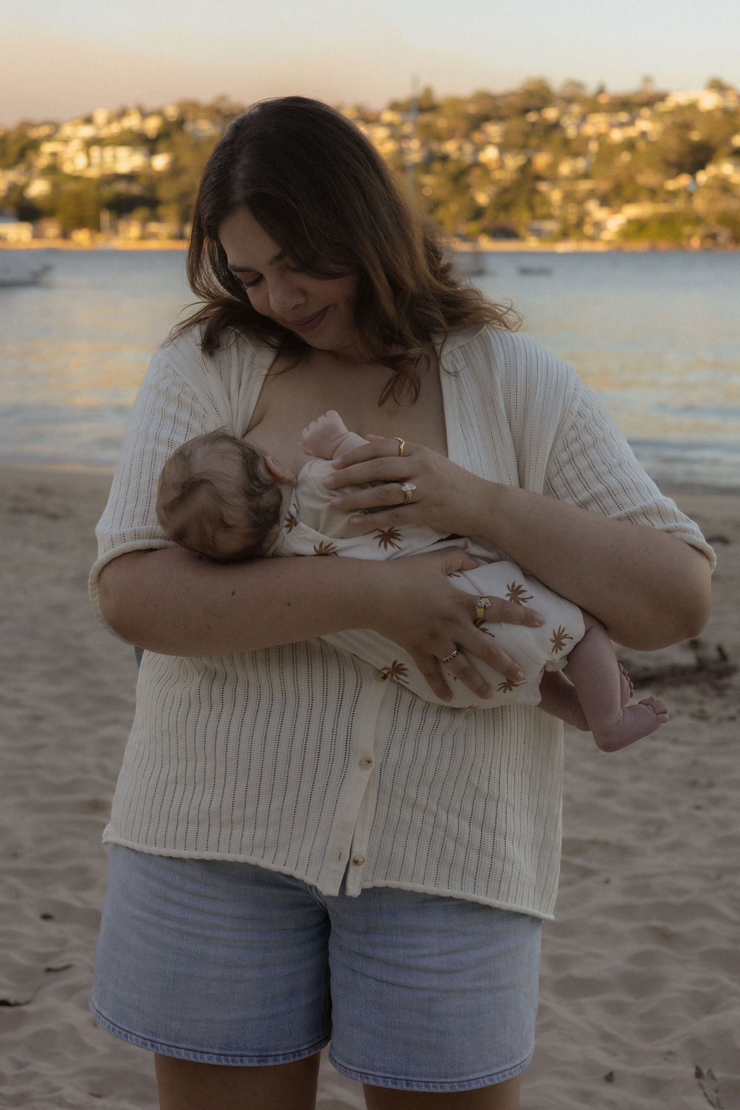 Mother breastfeeding newborn baby on sandy beach at sunset with waterfront suburb in background.