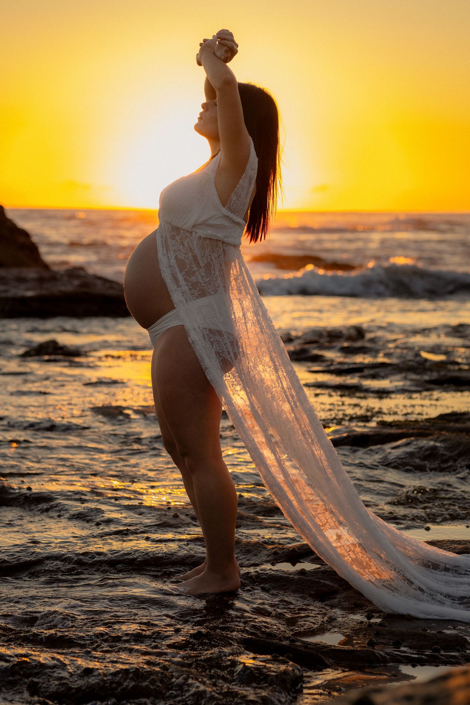 Pregnant woman in white lace robe poses gracefully on rocky beach at golden sunset with ocean waves behind her.