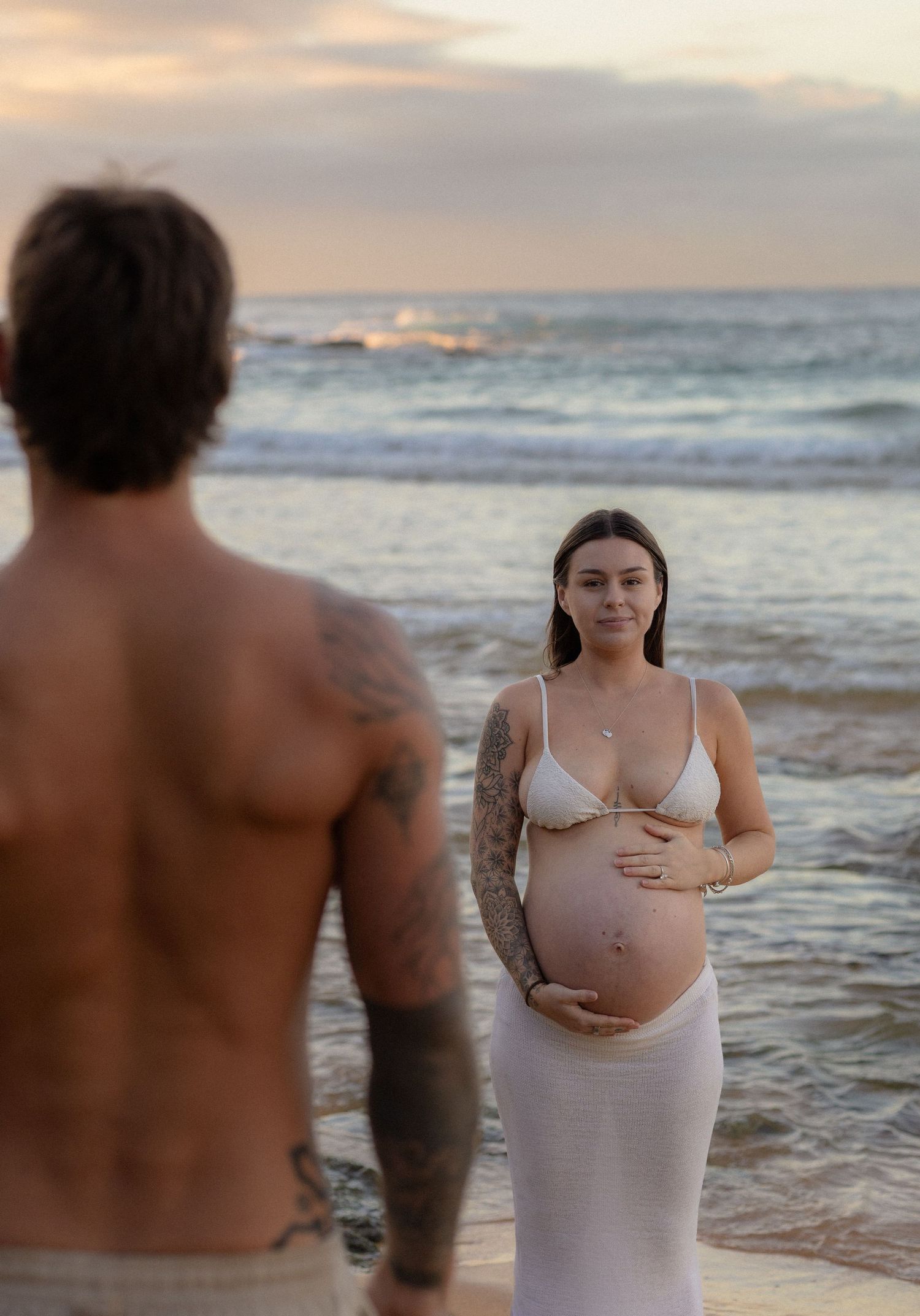 Pregnant woman in white bikini cradling baby bump on beach at sunset with tattooed partner.