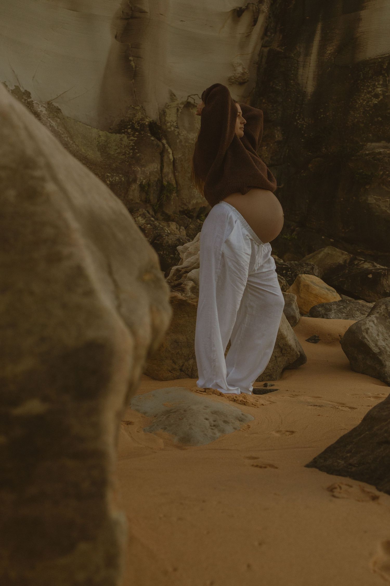 Pregnant woman in white pants cradling bare belly among rocky beach landscape in warm golden tones.