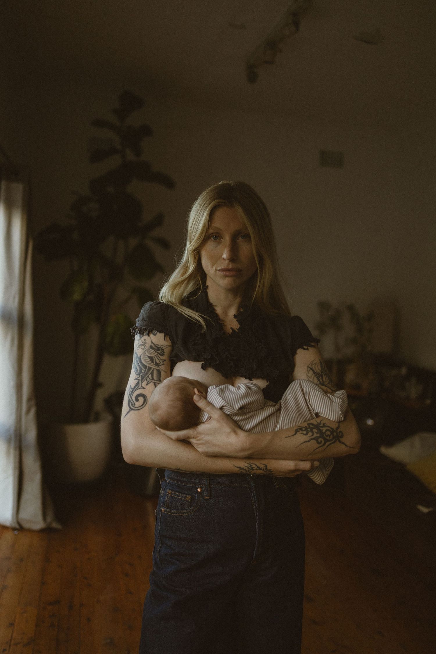 Tattooed blonde woman holding a newborn baby in a moody, plant-filled living room with hardwood floors.