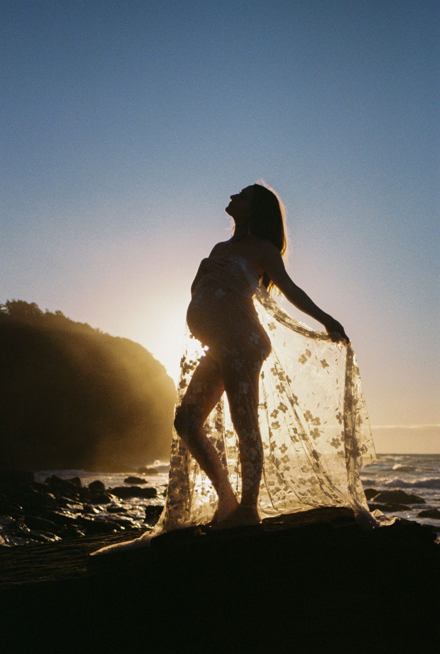 Silhouette of a pregnant woman holding sheer floral fabric at sunset on a rocky beach.