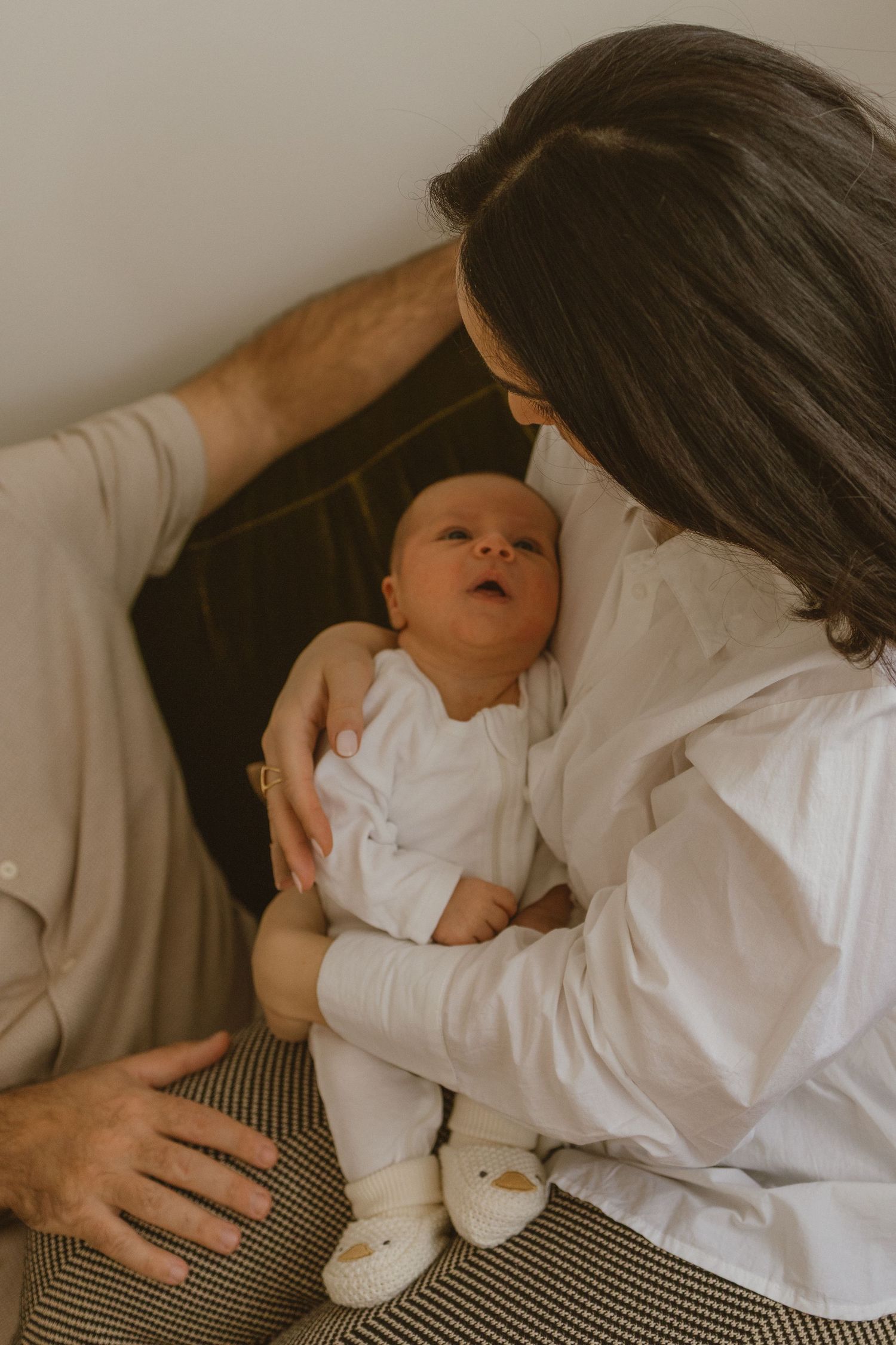 A woman in a white shirt holds a newborn baby in white clothing while a man gently places his hand on the infant.