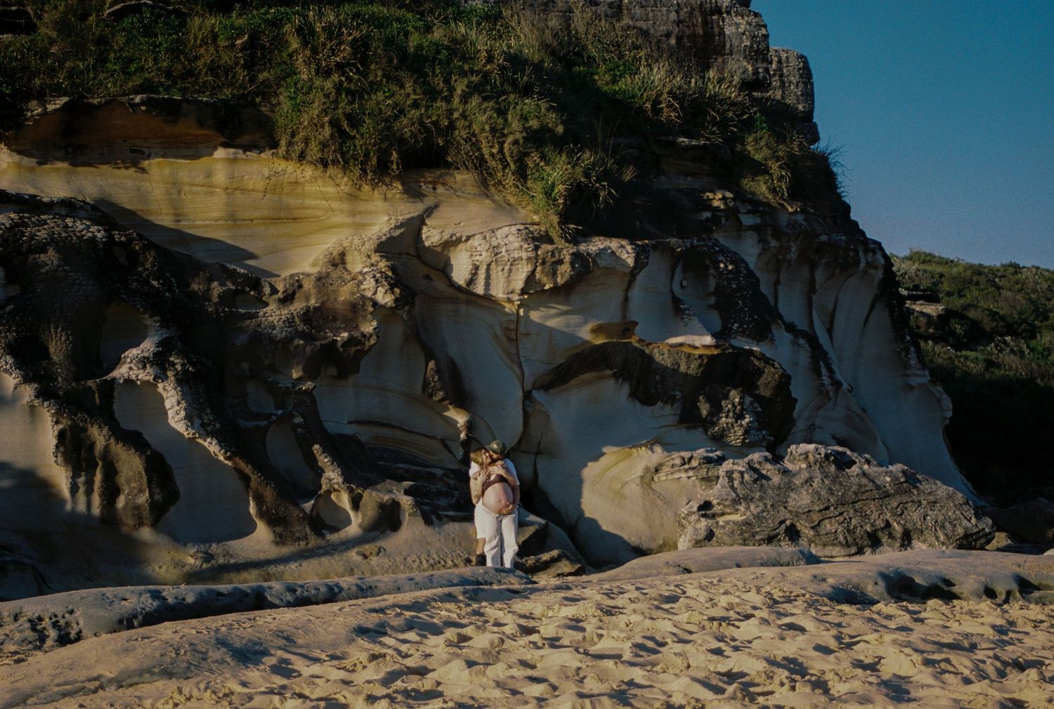 Couple embracing on sandy beach beside dramatic eroded sandstone cliffs with lush green vegetation above.