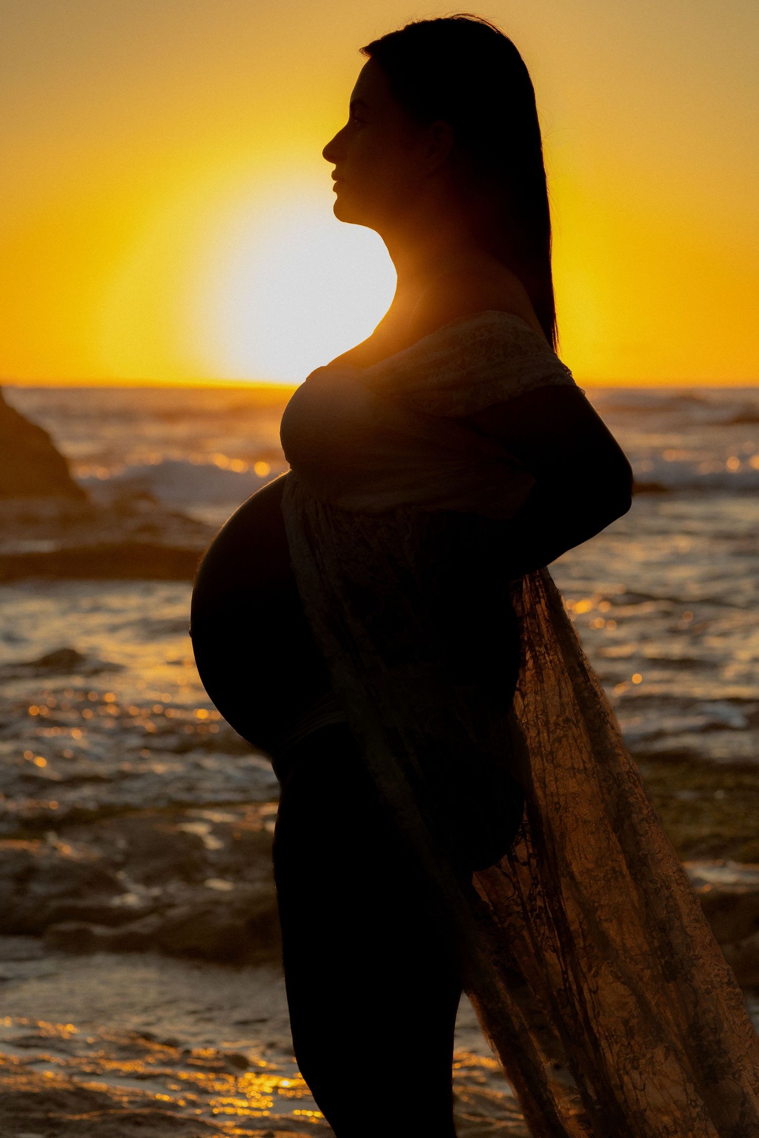 Silhouette of a pregnant woman standing on a rocky beach at golden sunset with ocean waves in the background.