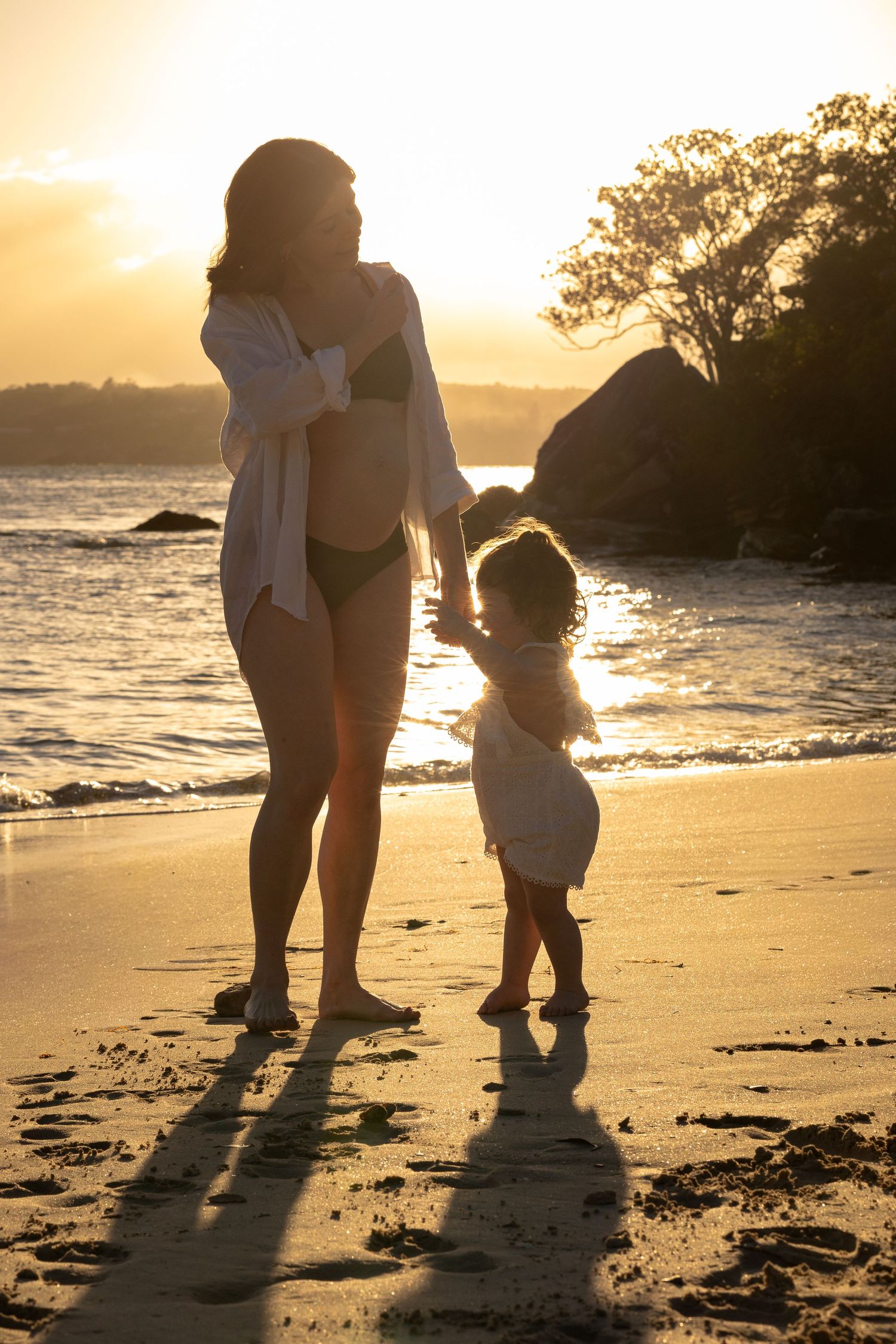 A woman in a white cover-up stands on a beach at sunset, with the ocean in the background.