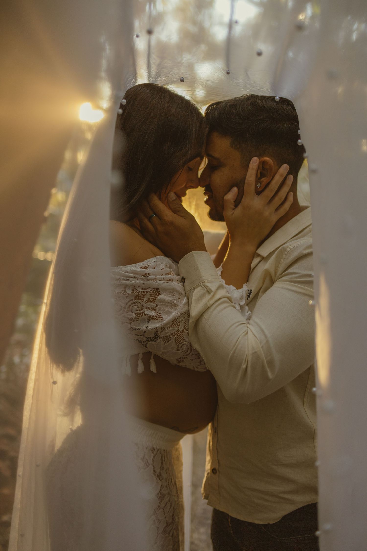 Romantic couple embracing tenderly with foreheads touching, surrounded by warm fairy lights and sheer curtains.