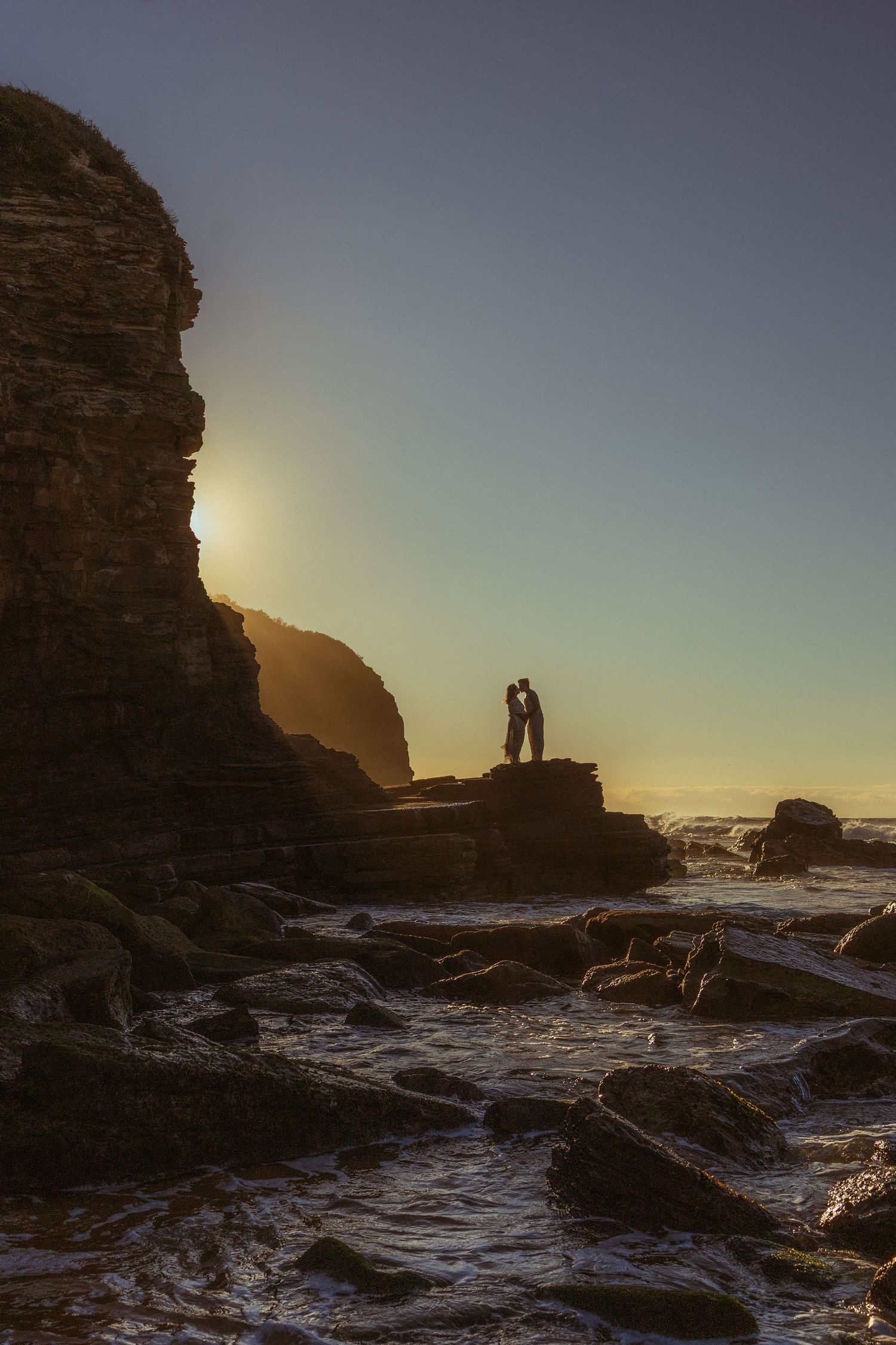 Silhouette of couple standing on rocky coastal cliffs at golden sunset with ocean waves below.