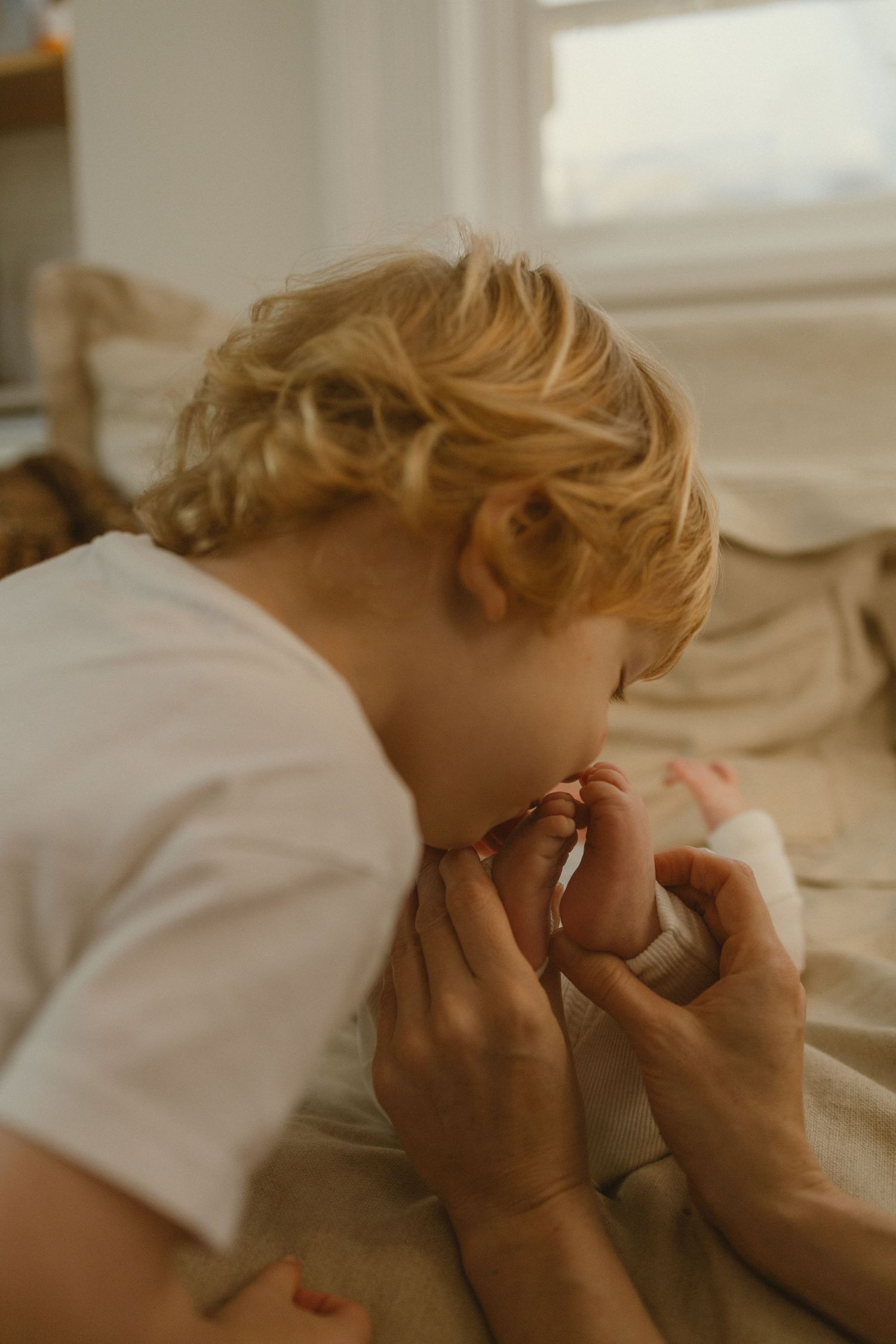 Blonde toddler kissing tiny newborn baby feet held by an adult in a cozy room.