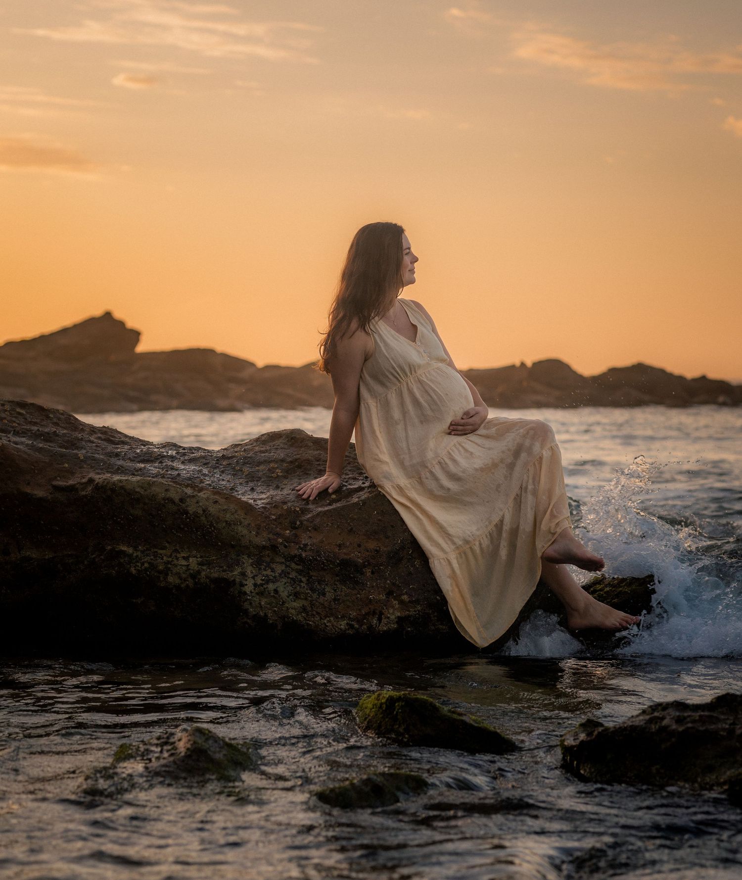 A figure in a flowing white dress sits on coastal rocks at sunset while ocean waves crash around them.