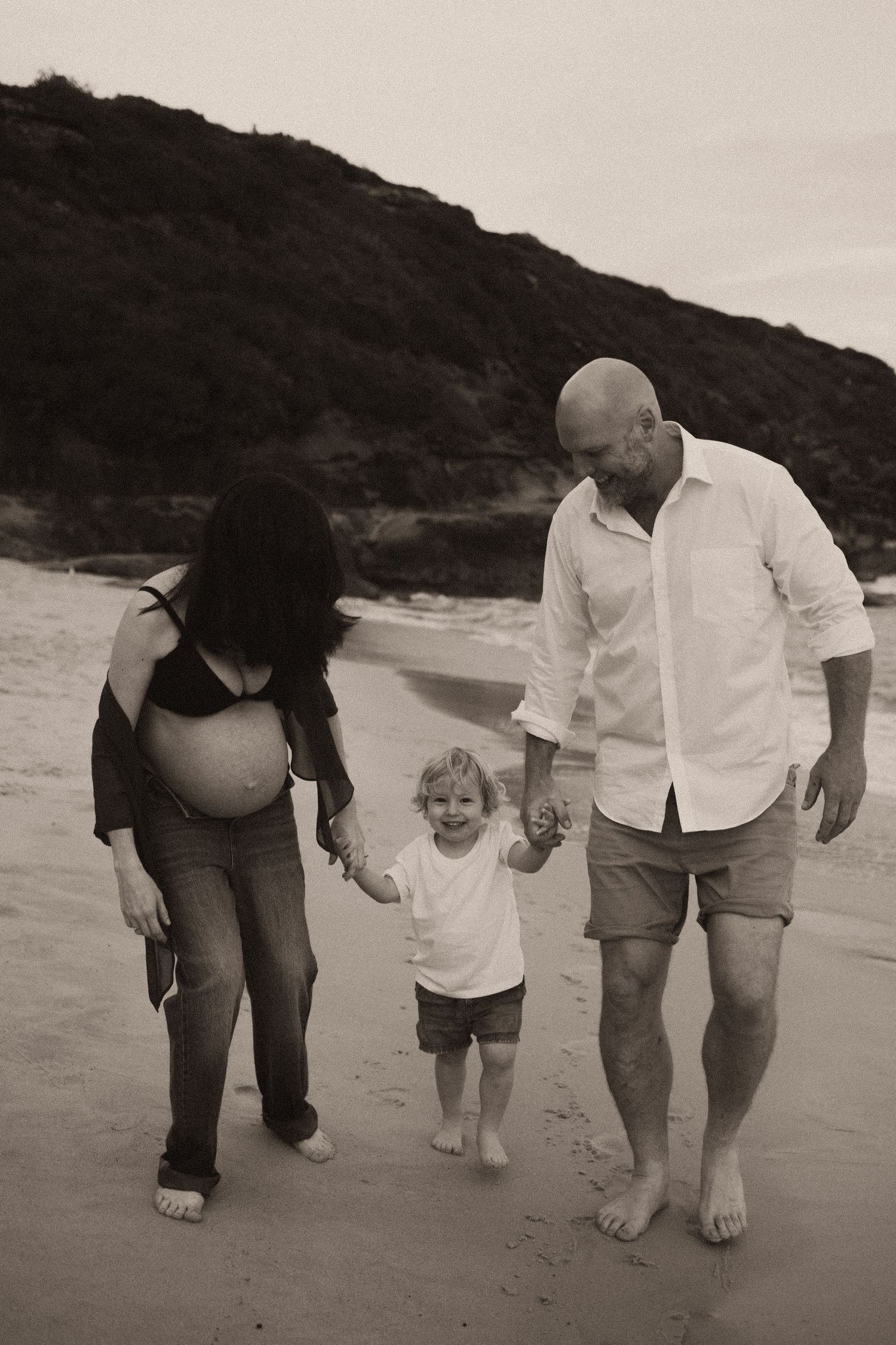 Pregnant woman and bald man walk hand-in-hand with toddler on beach in sepia-toned family maternity photo.