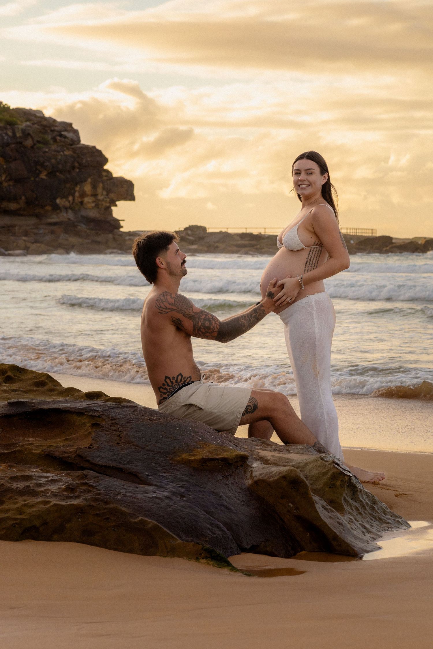 Shirtless tattooed man kneels before smiling pregnant woman on beach rocks at golden sunset.
