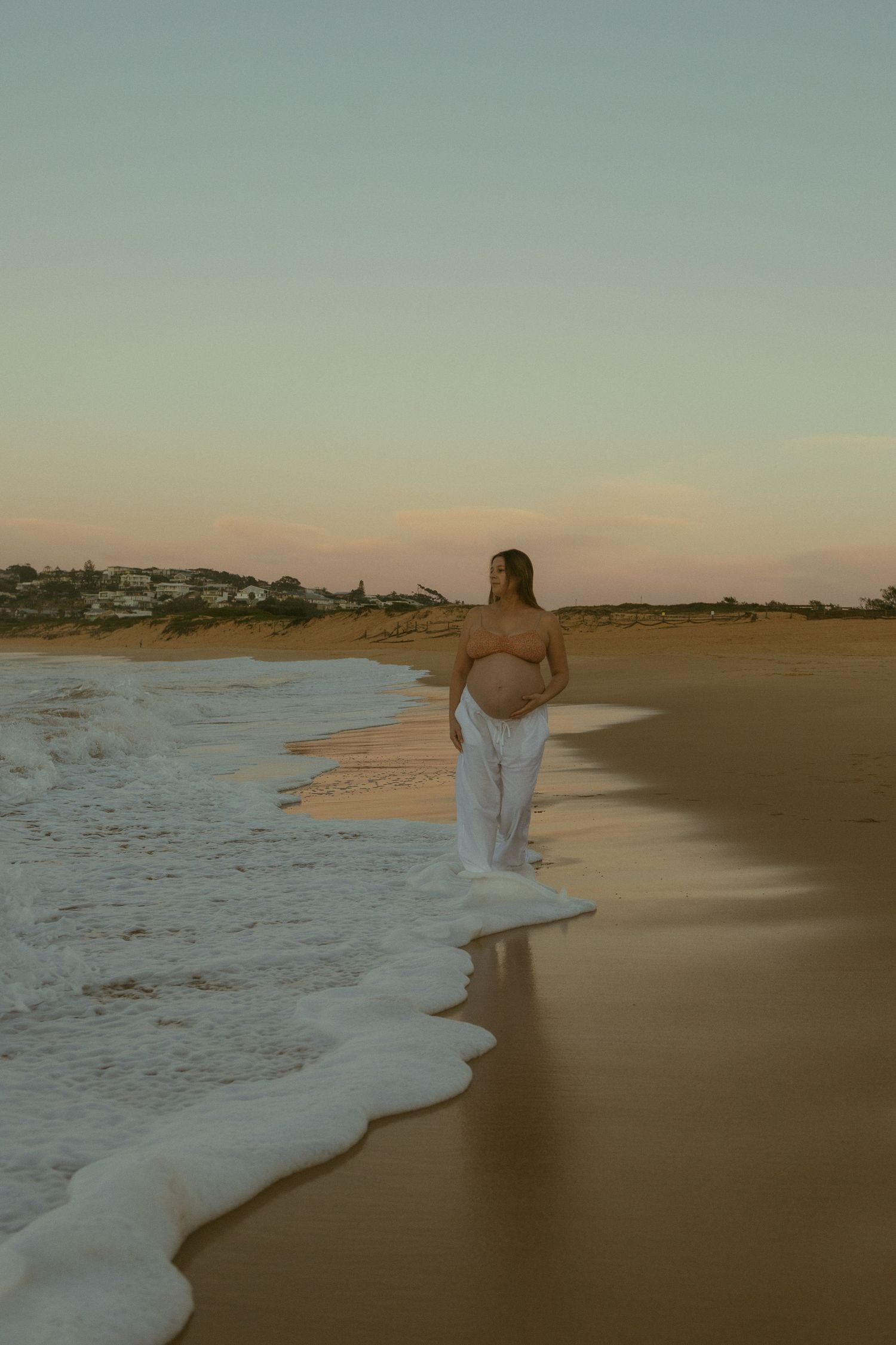 Pregnant woman standing on sandy beach at sunset, wearing white pants, with ocean waves at her feet.