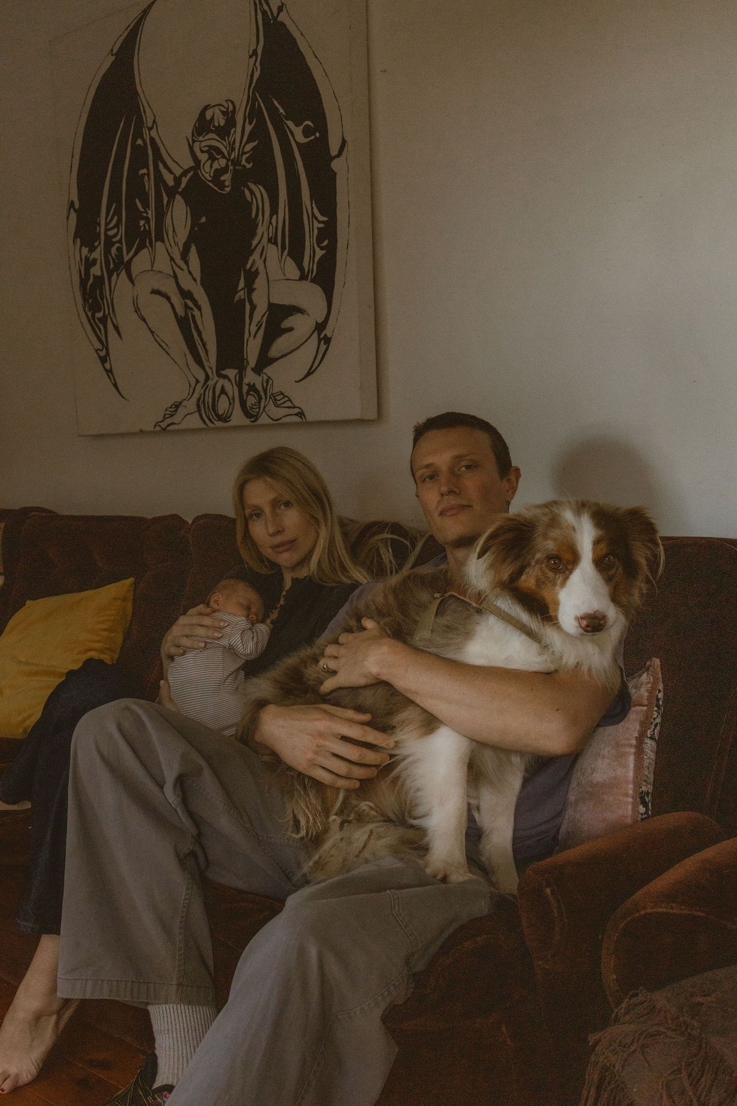 Couple sitting on brown sofa holding large Australian Shepherd dog and baby, with dark fantasy artwork on wall.