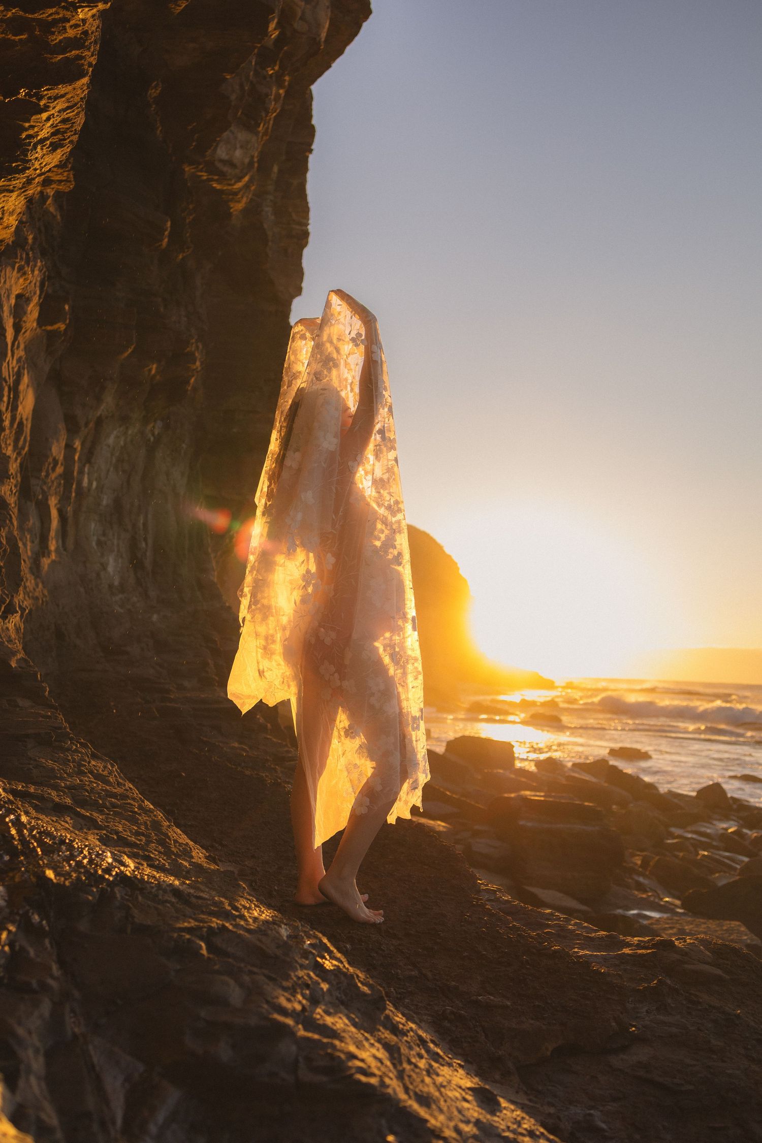 Pregnant woman draped in sheer white fabric backlit by stunning golden sunset on rocky shore.