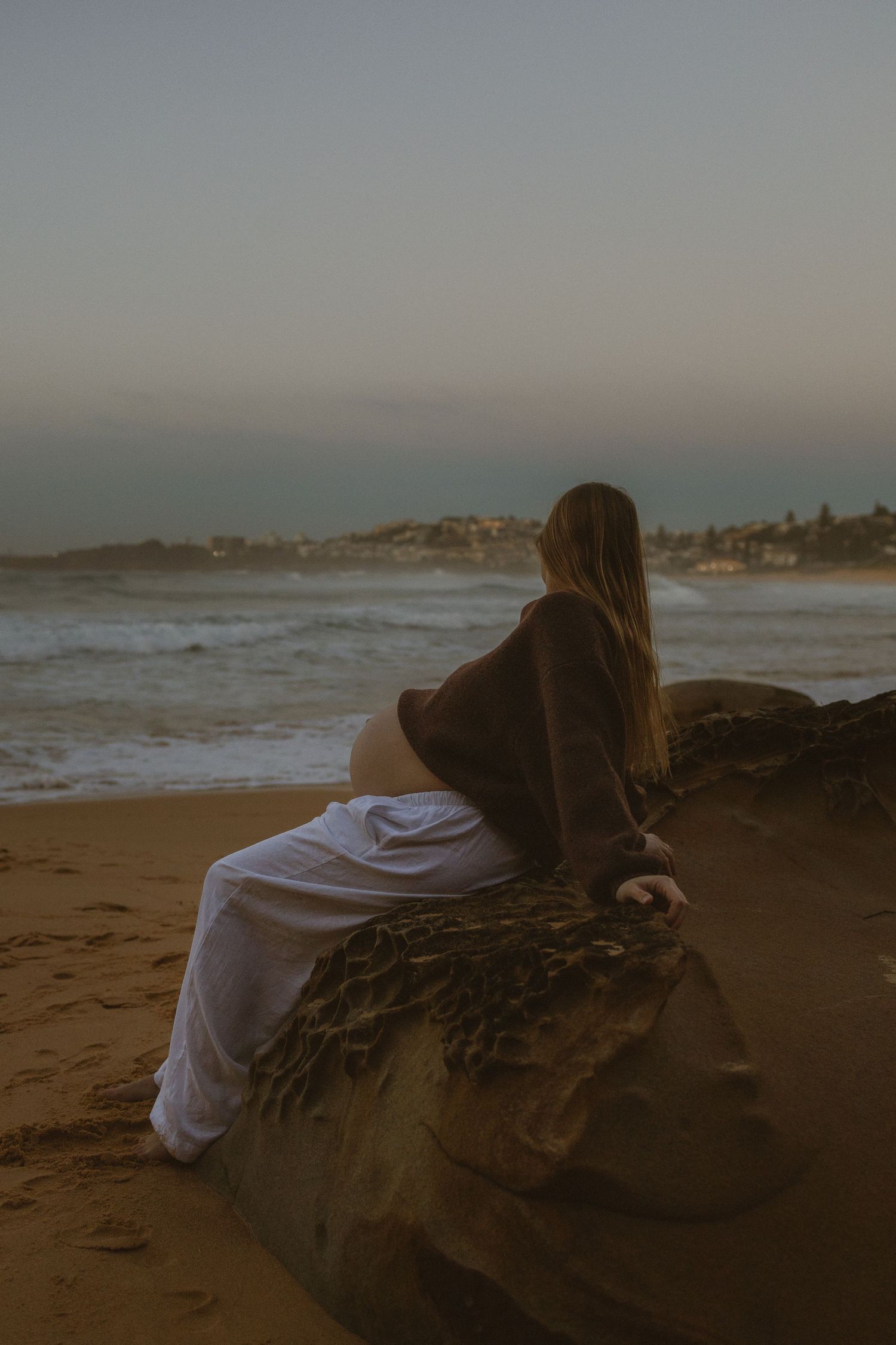 Woman in white pants and brown sweater sits on a rock gazing at ocean waves at dusk on a sandy beach.