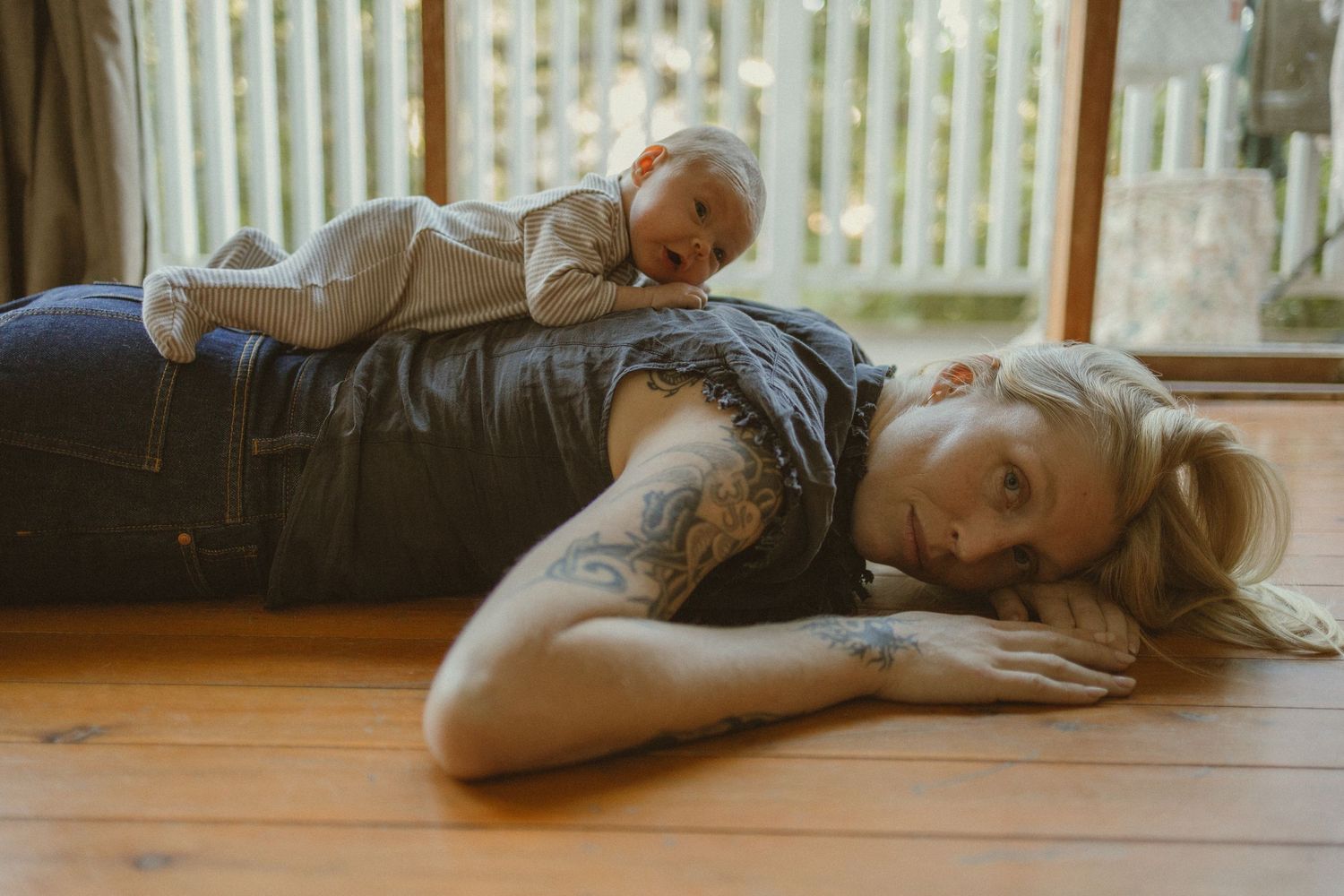 Tattooed mother lying on hardwood floor while baby rests on her back in a cozy indoor setting.
