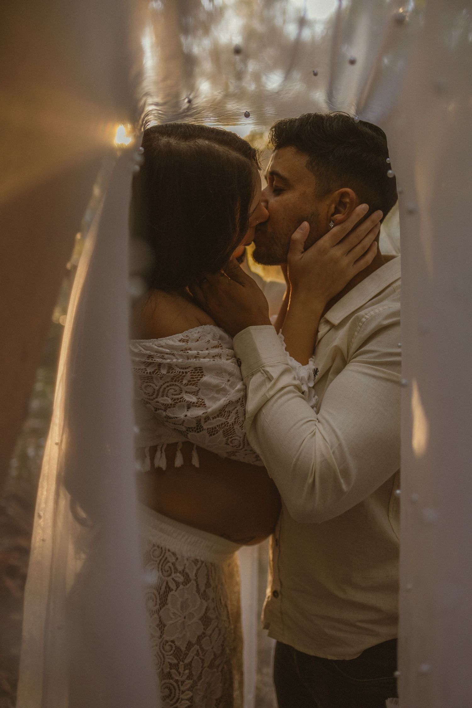 Couple sharing a romantic kiss framed by white drapes with warm bokeh lights in the background.