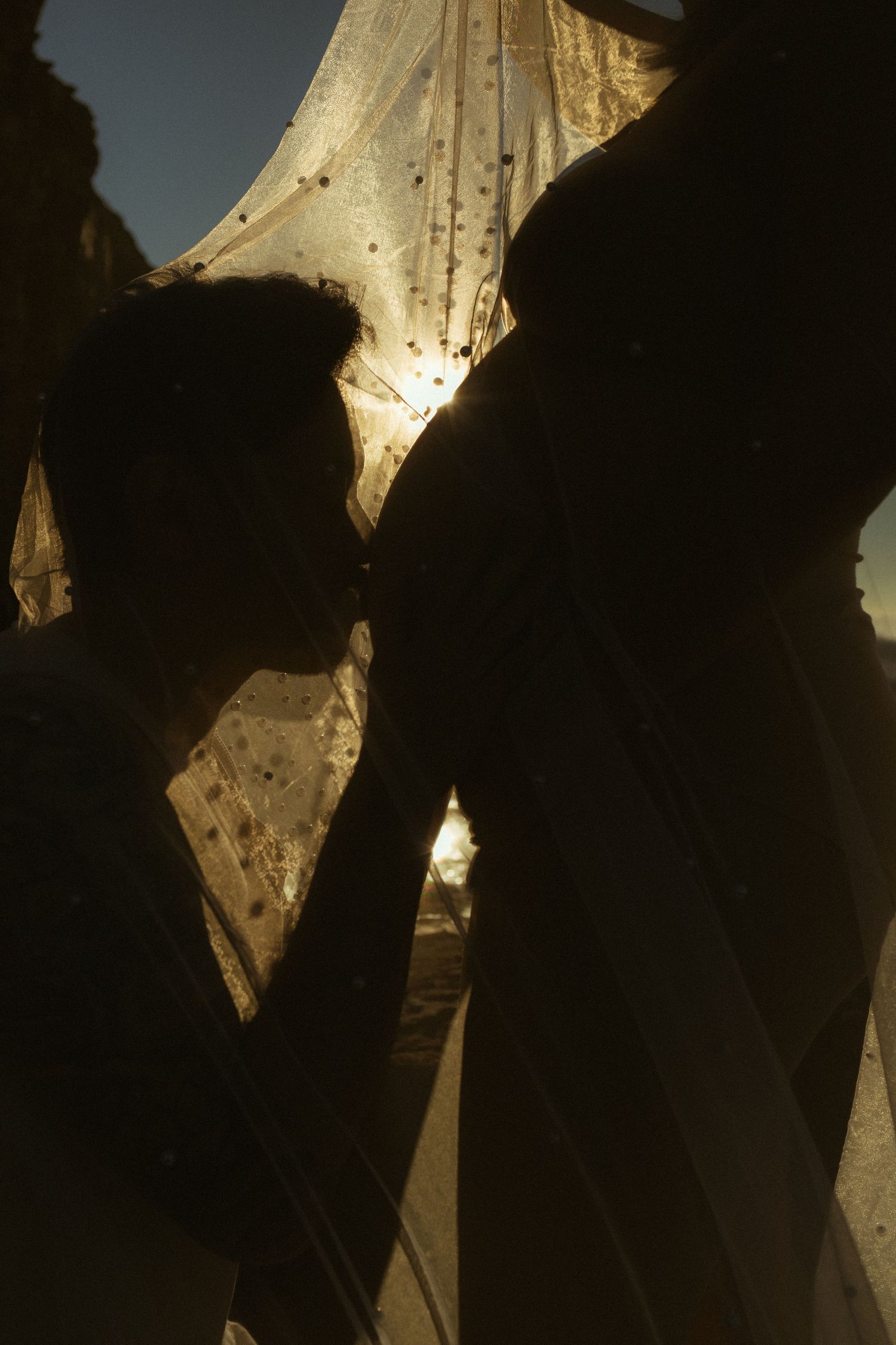 Silhouette of couple kissing under golden backlit wedding veil at sunset, romantic bridal portrait.
