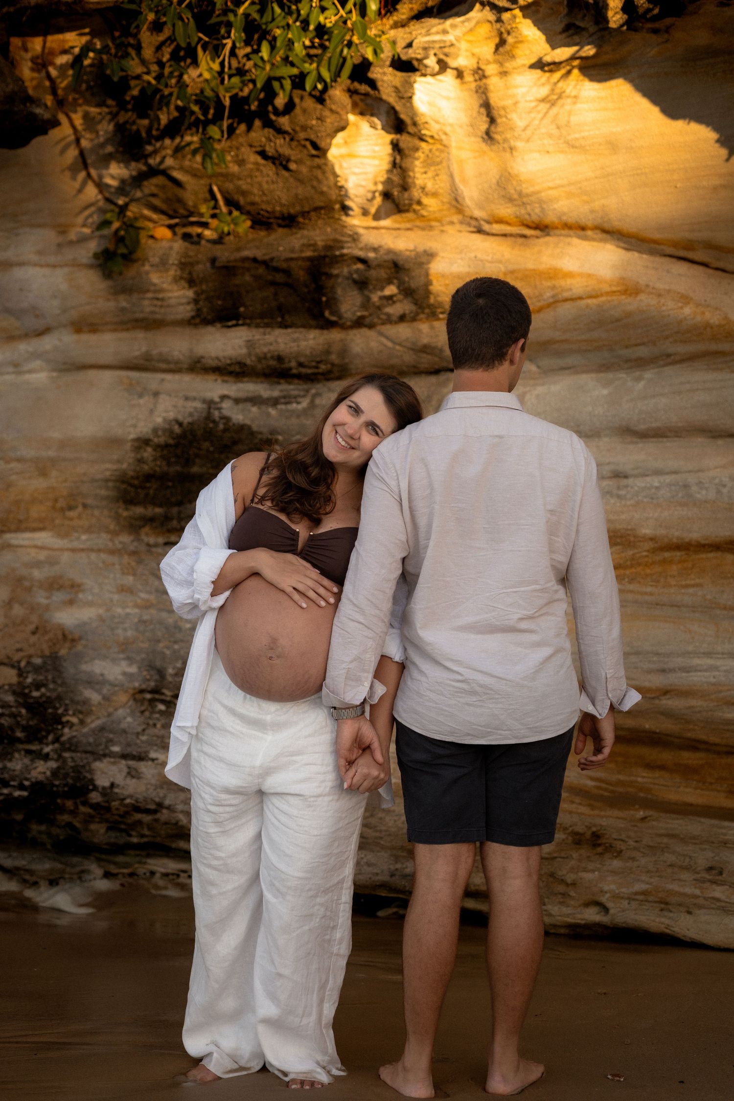 A couple stands on a beach at sunset with dramatic sandstone cliffs in the background creating a warm, glowing atmosphere.