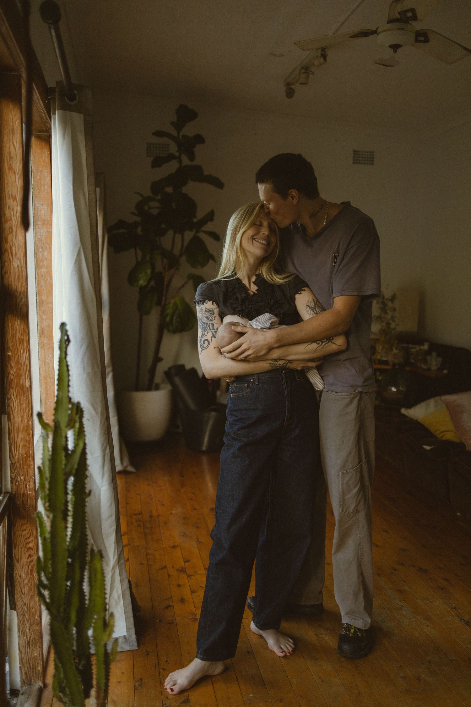 A couple stands barefoot indoors holding their newborn baby, surrounded by houseplants in a warm, cozy living room.