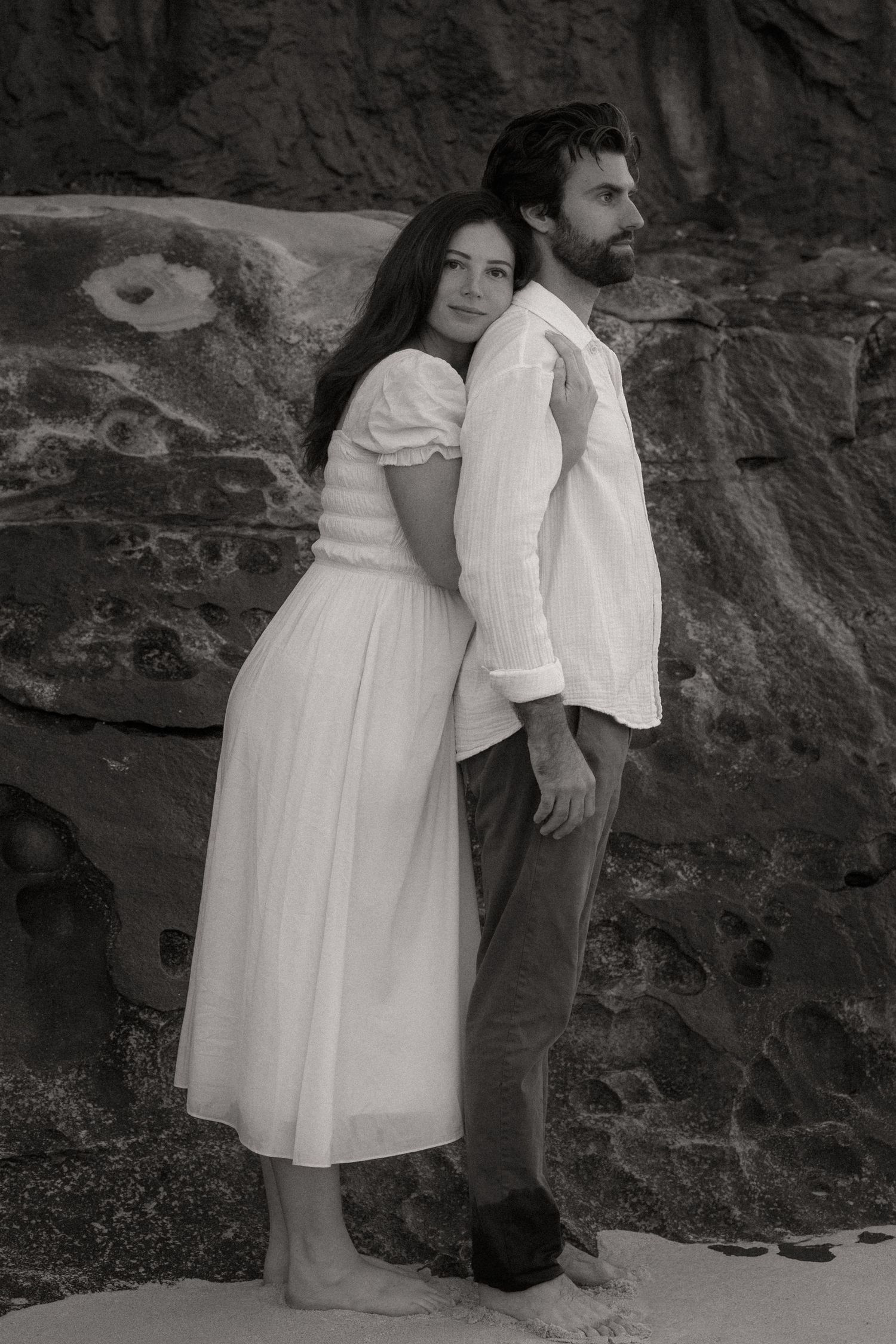 Black and white photo of a couple posing against rocky cliffs, woman in white dress embracing man.