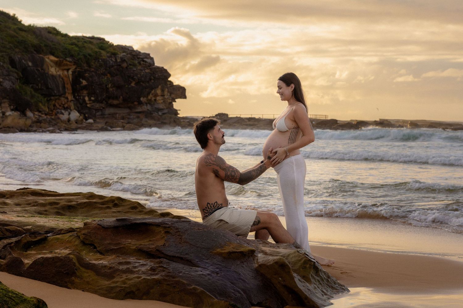Couple sharing tender maternity moment on rocky beach at golden sunset, man kneeling before pregnant woman.