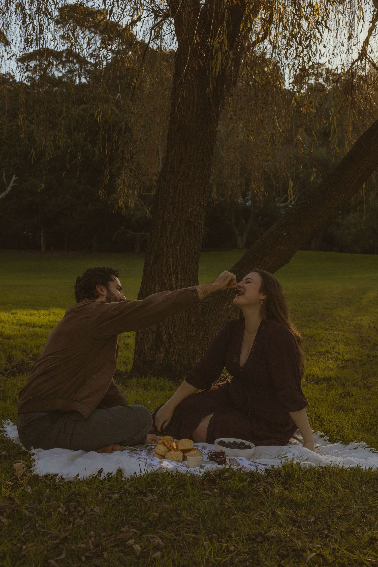 Couple sharing a romantic picnic under a willow tree at sunset, man feeding woman fruit on a white blanket.