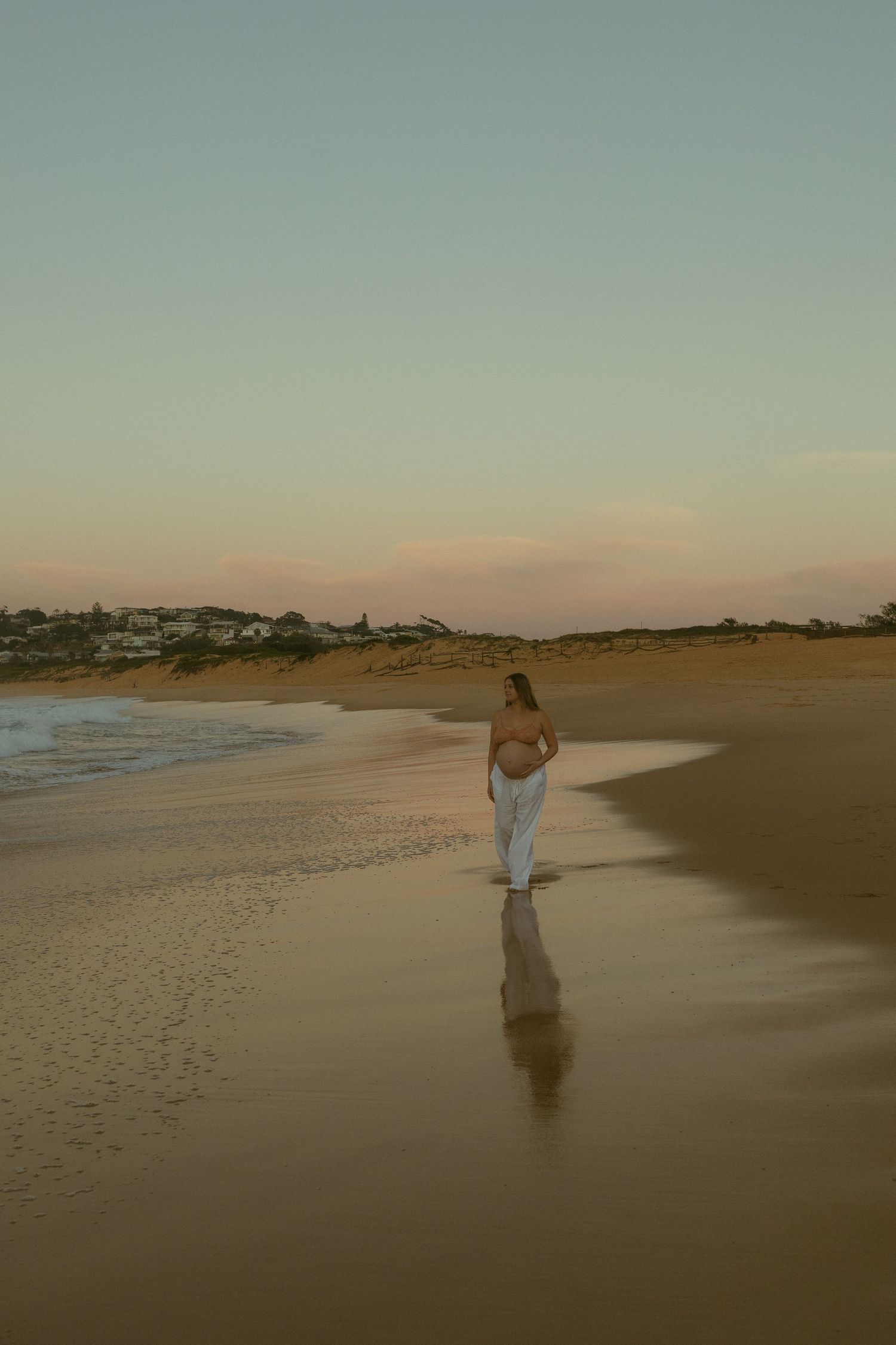 Pregnant woman in white pants walks along a sandy beach at sunset with gentle waves and coastal scenery.