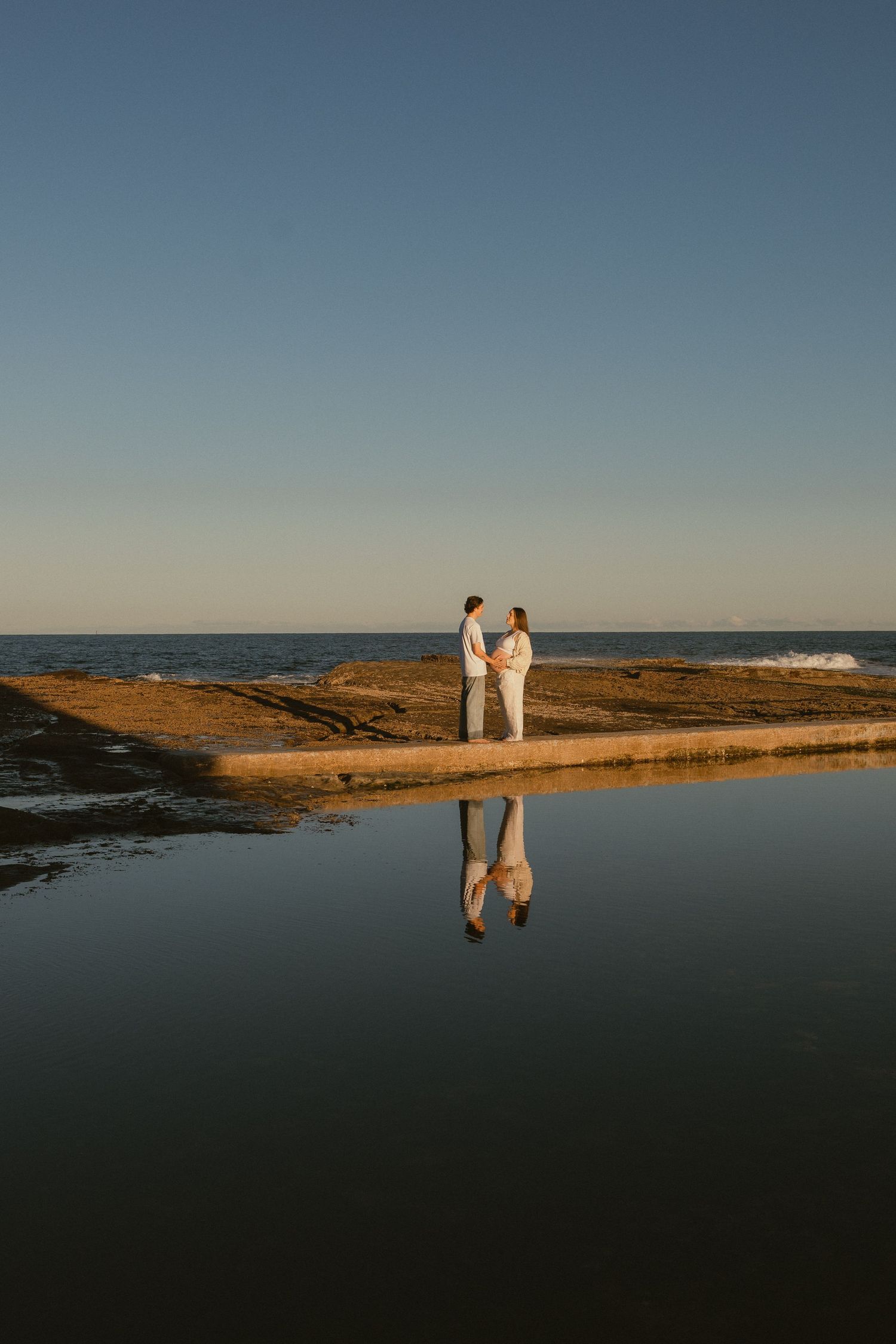 Couple holding hands on a rocky coastal platform at golden hour, with their reflection mirrored in calm water below.
