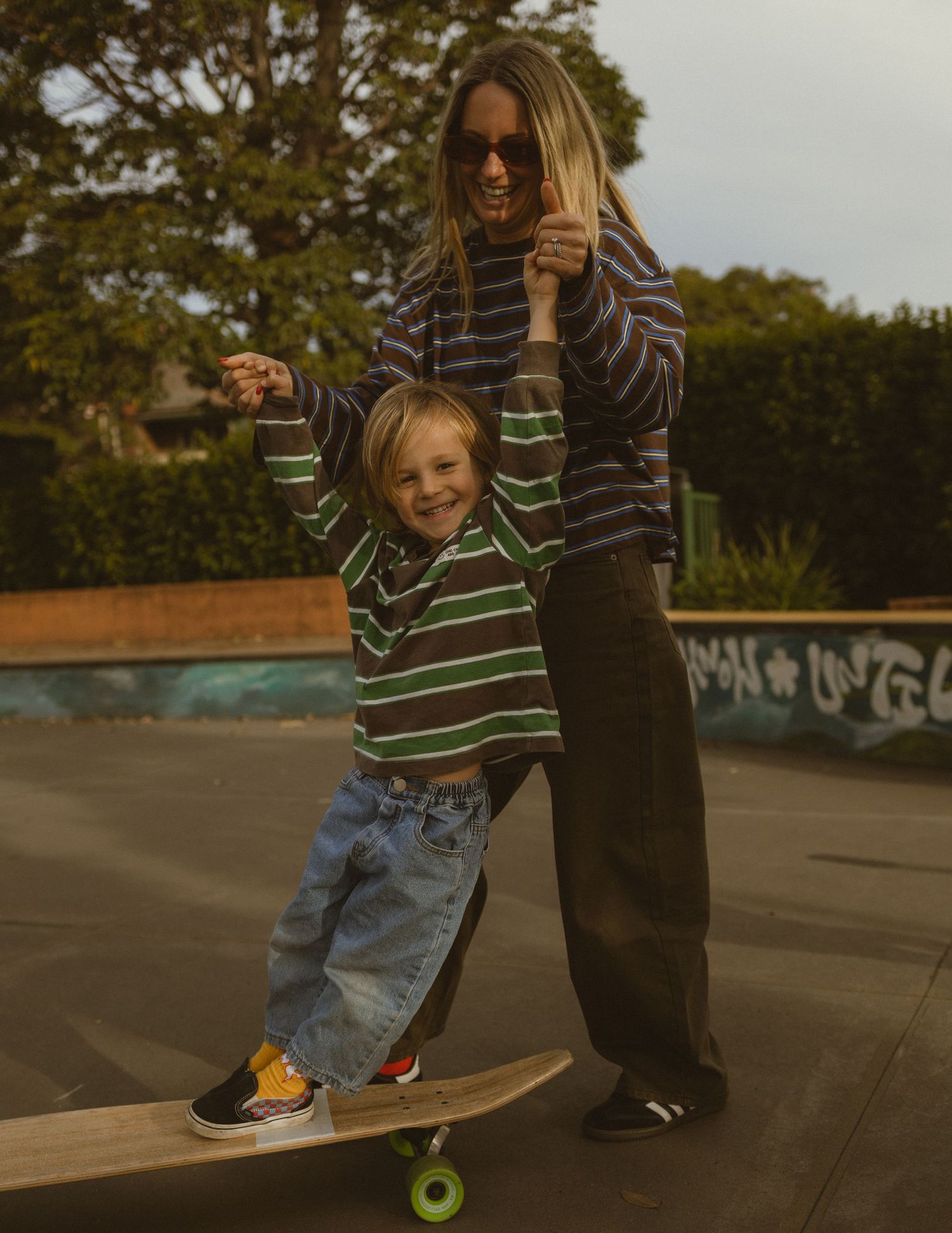 A woman helps a young child ride a skateboard at a skate park, both wearing striped tops and smiling.