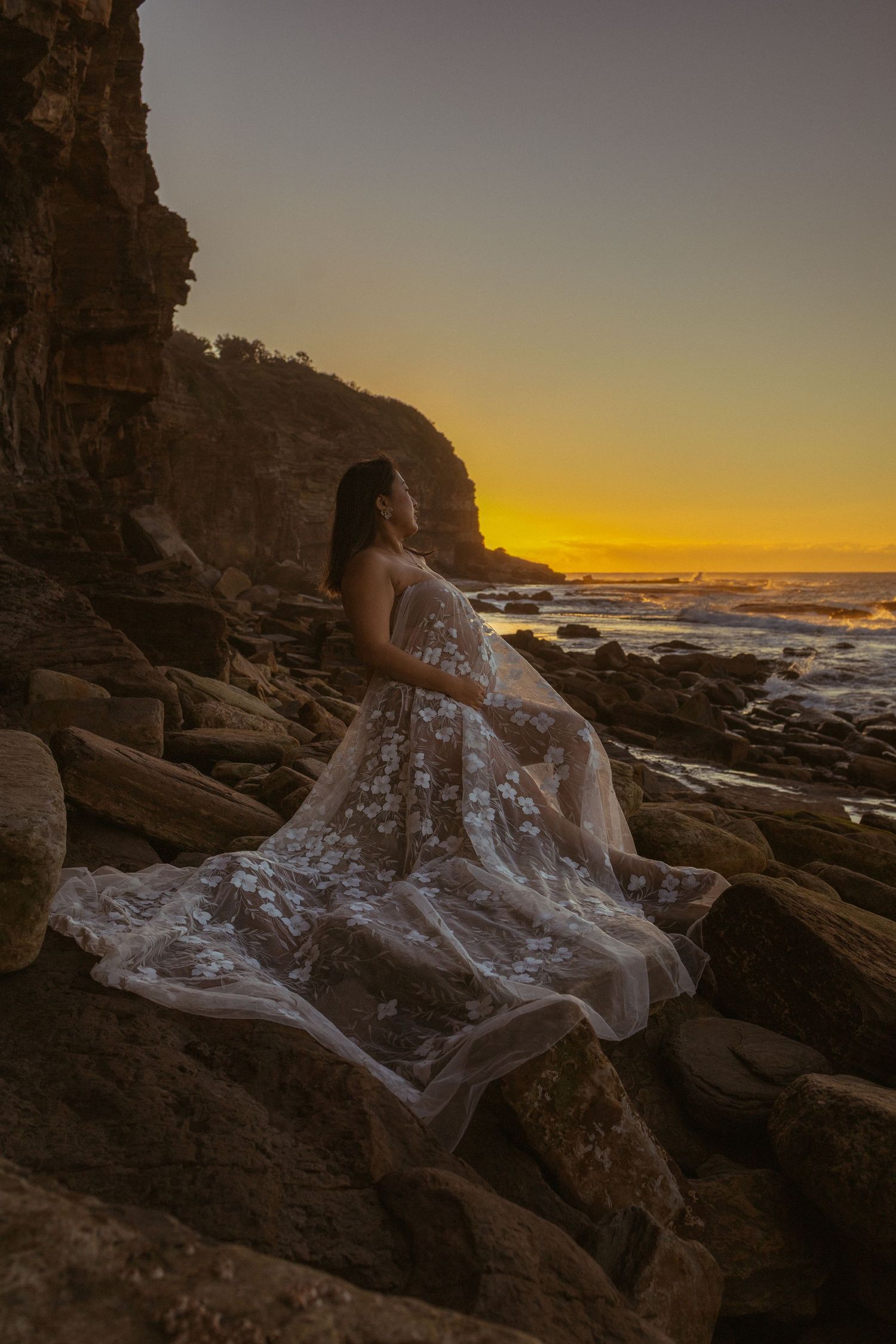 Woman in flowing white lace gown poses on rocky coastline at golden sunset with ocean waves in background.