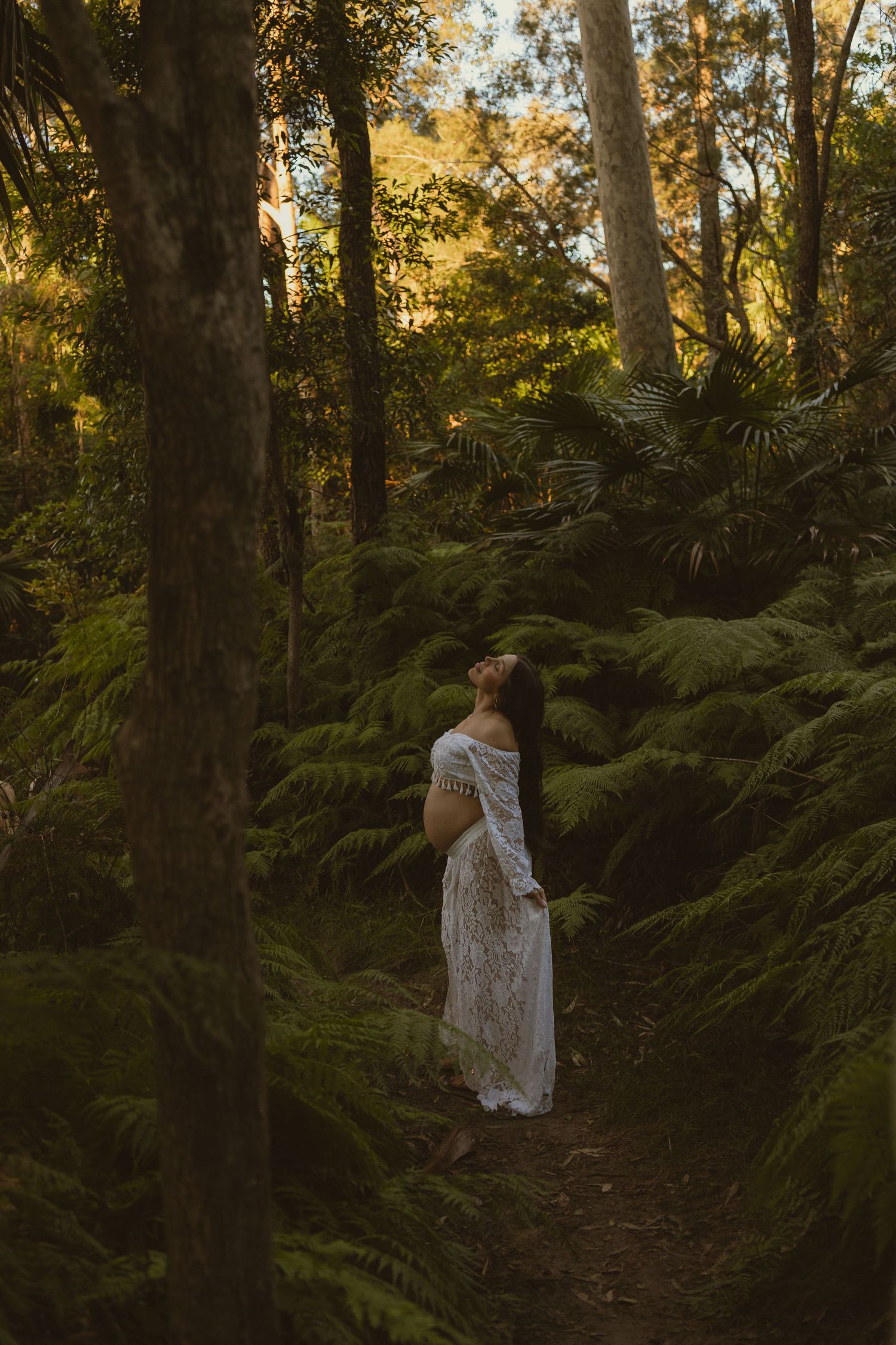 Expectant mother in white lace gown posing gracefully on a forest path surrounded by tree ferns.
