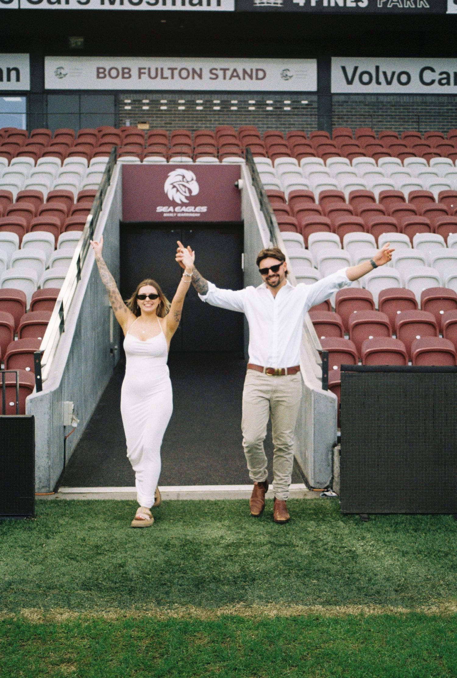 Couple in white outfits celebrating at Bob Fulton Stand, Manly Sea Eagles stadium, raising arms joyfully.