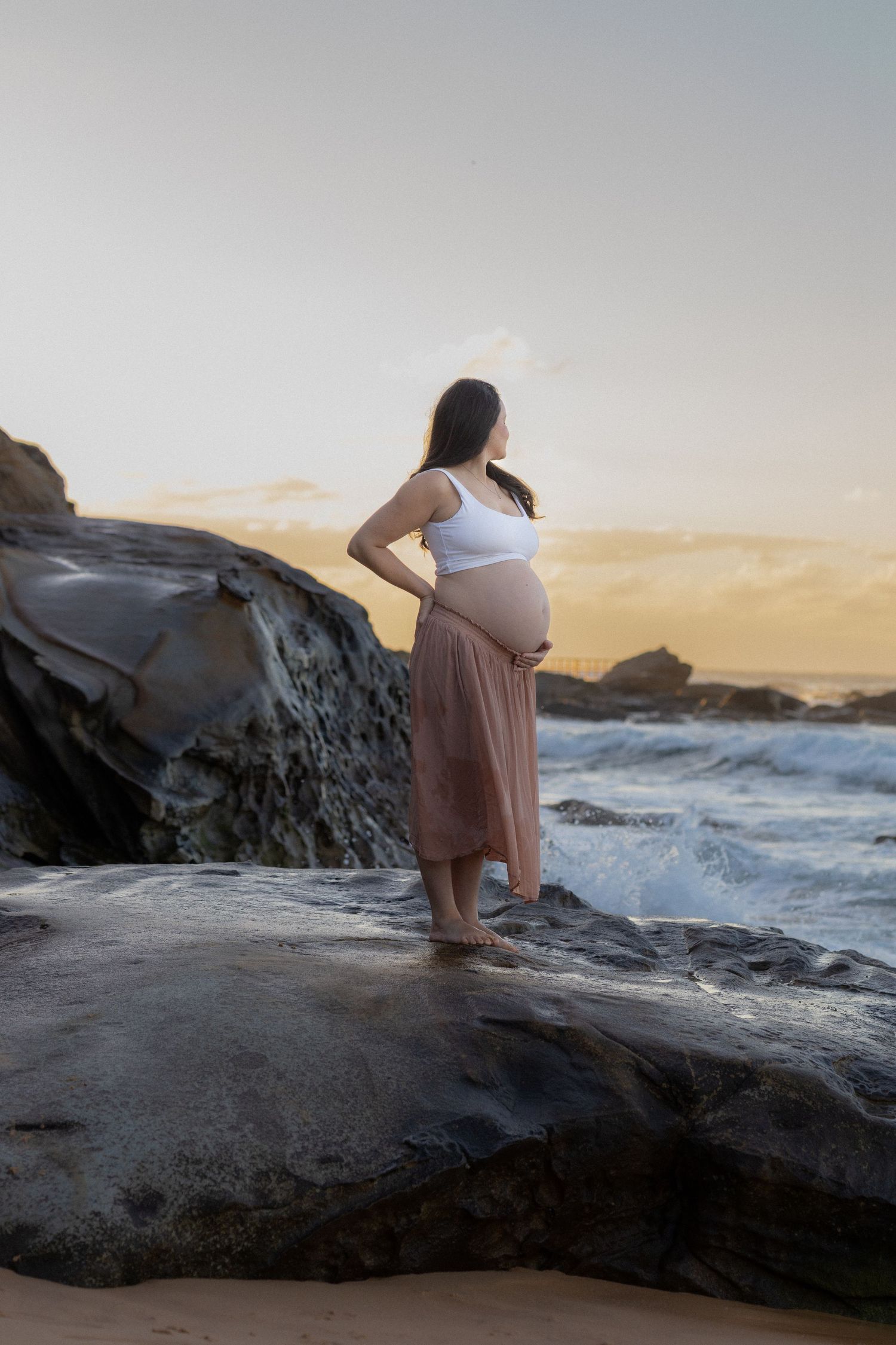 Expectant mother in flowing skirt stands on coastal rocks at sunset while waves crash against the shore.