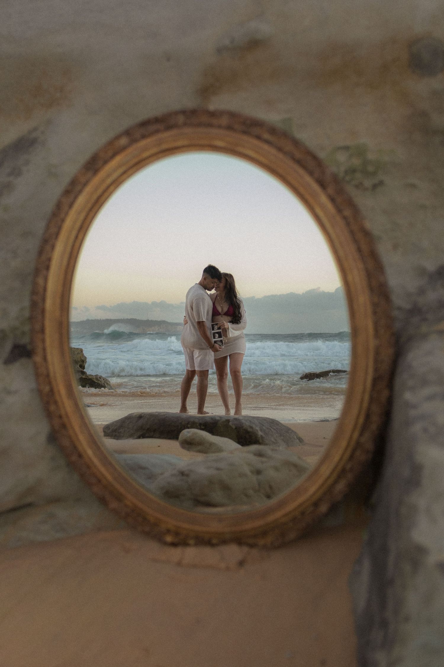 Couple kissing on a rocky beach at sunset reflected in an ornate oval gold mirror.